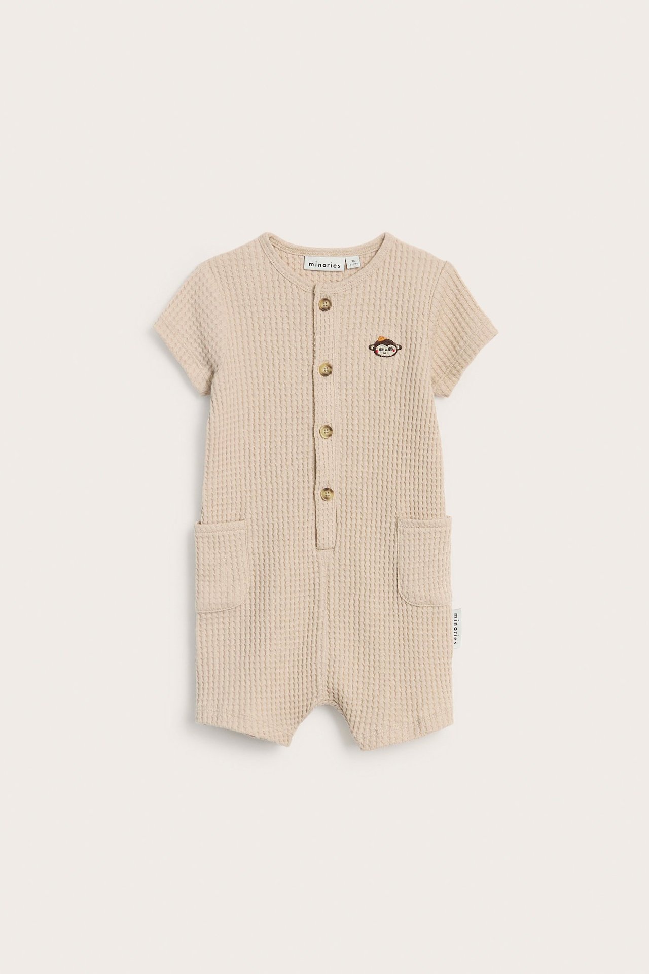 Edestä näkymä beigestä vohvelikankaasta vauvan haalarista, jossa napit edessä, sivutaskut ja apinakirjonta.