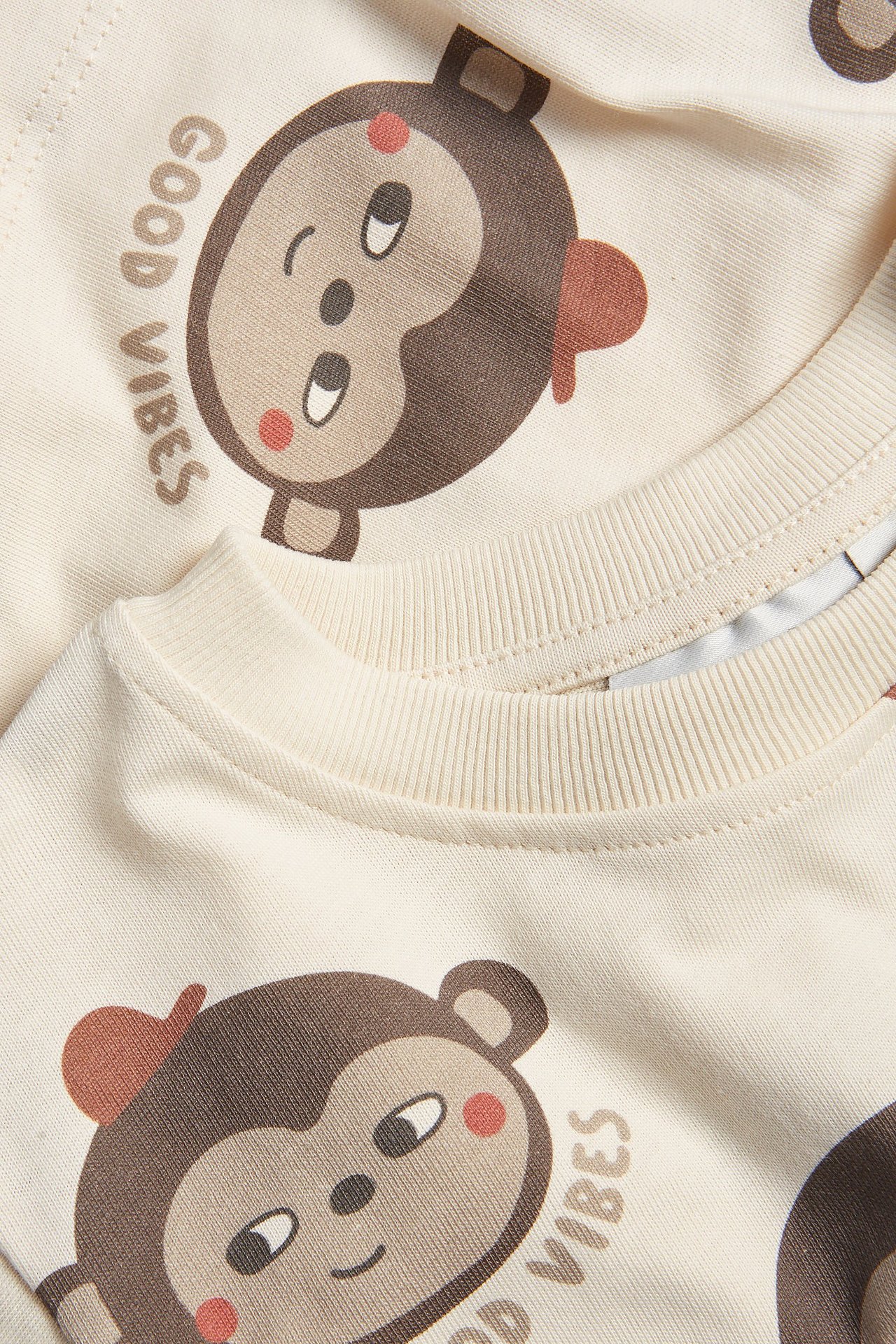 Lähikuva kahdesta vaaleanbeigestä puuvillaisesta vauvan t-paidasta, joissa apinaprintti ja 'GOOD VIBES' -teksti.