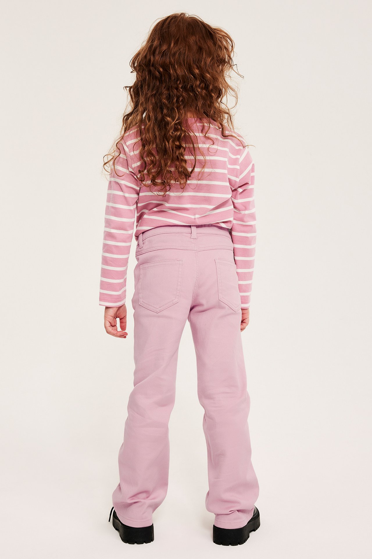 Ryggvy av ett barn i ljusrosa bootcut jeans med medelhög midja och en rosa-vit randig långärmad tröja.