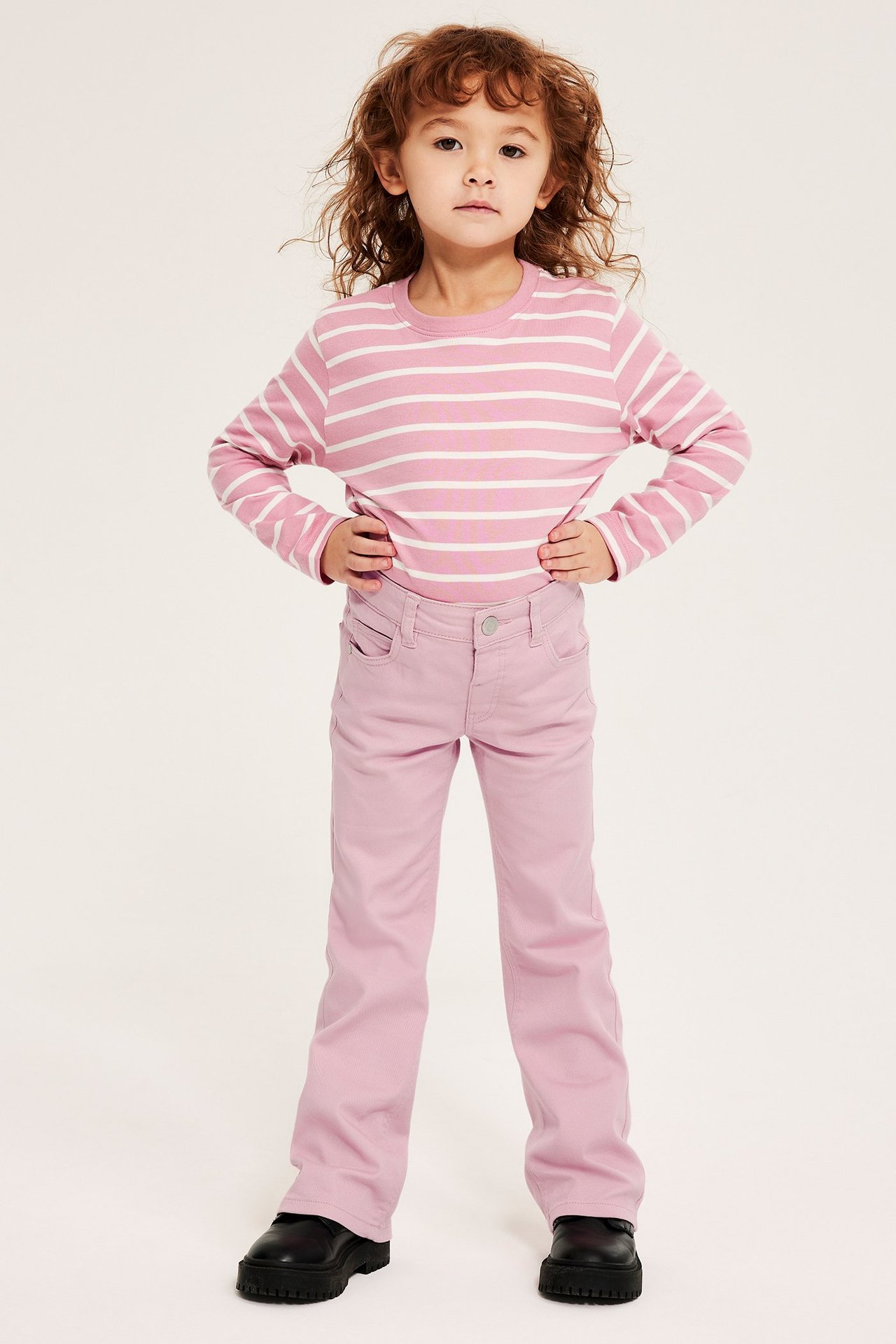 Framsida: barn i ljusrosa bootcut-jeans, en rosa/vitrandig topp och svarta skor.