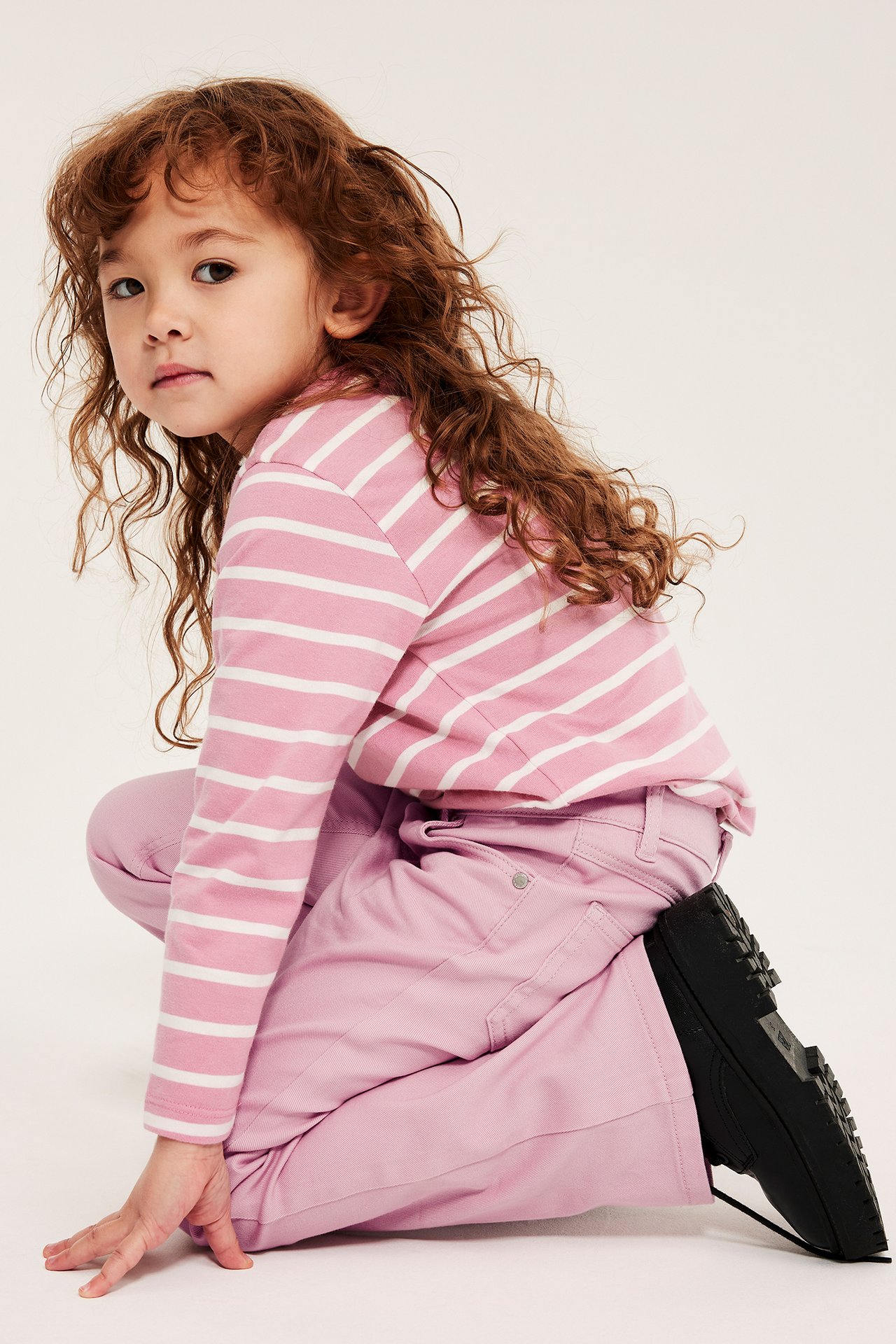 Sivukuva polvistuvasta lapsesta vaaleanpunaisissa bootcut-farkuissa, raidallisessa paidassa ja mustissa kengissä.