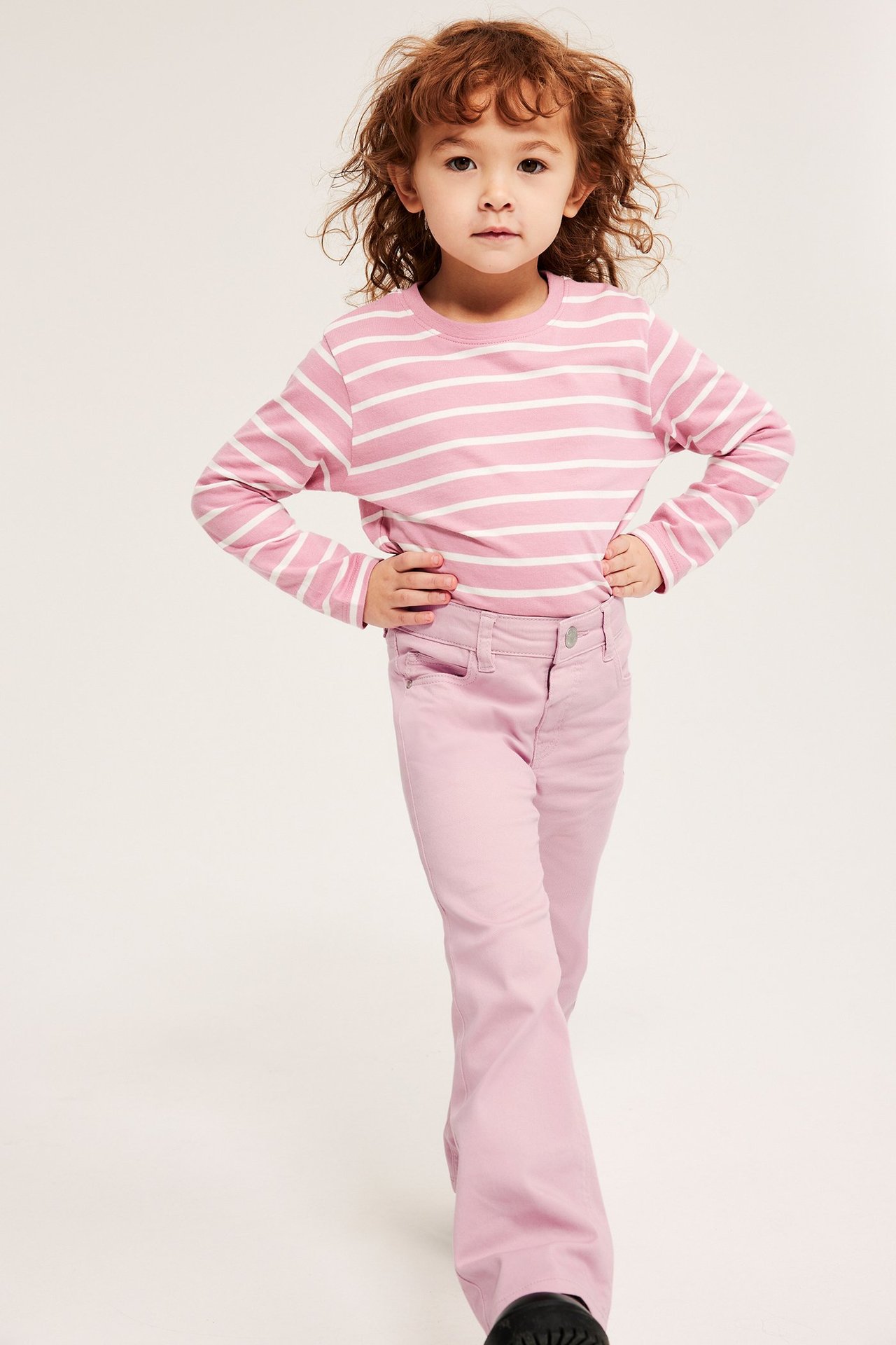 Framifrån, ett barn i ljusrosa bootcutjeans med medelhög midja och rosa- och vitrandig långärmad topp.