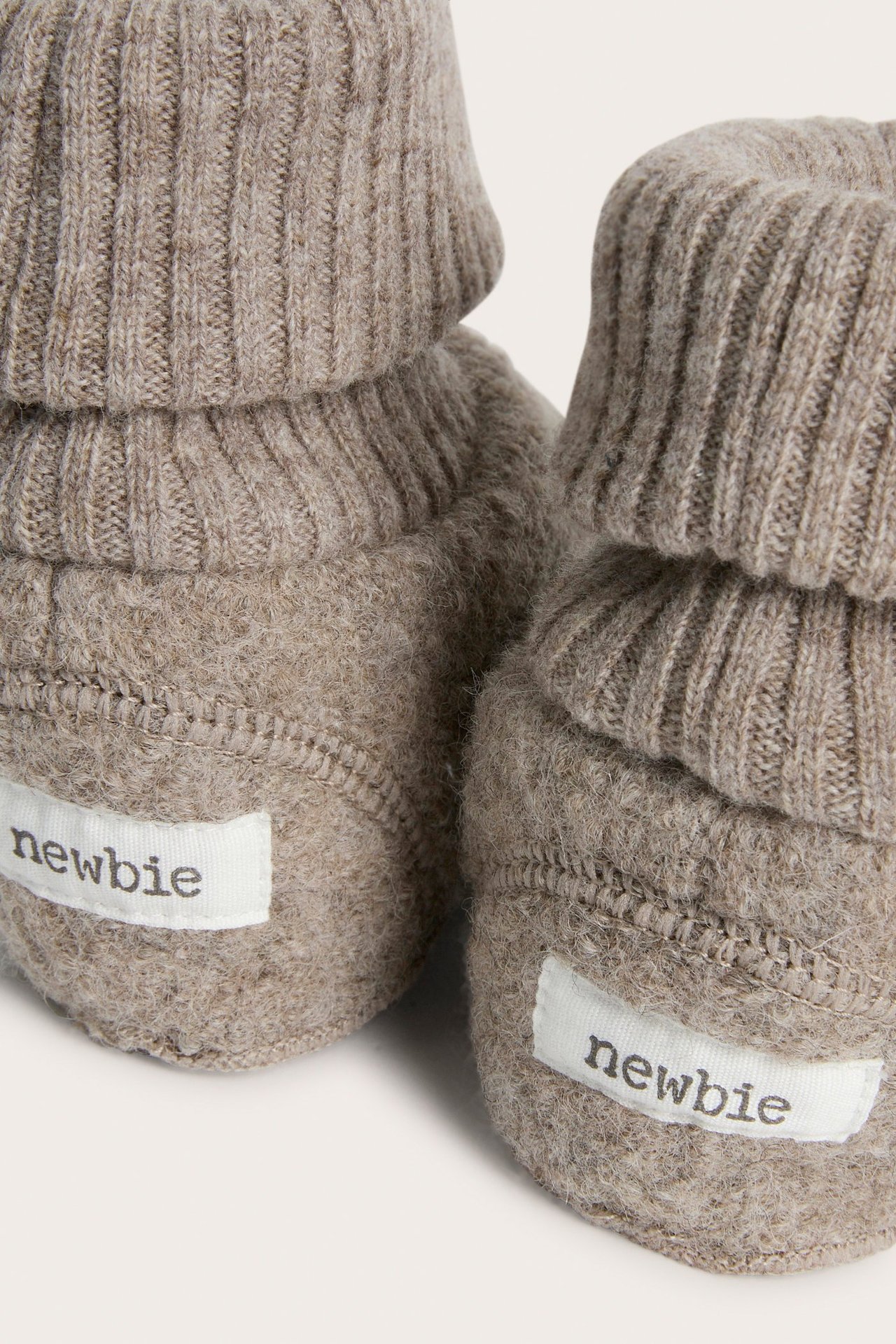 Nærbilde av to melert brune babyulltøfler med ribbestrikkede, nedbrettede mansjetter og hvite 'newbie'-etiketter.