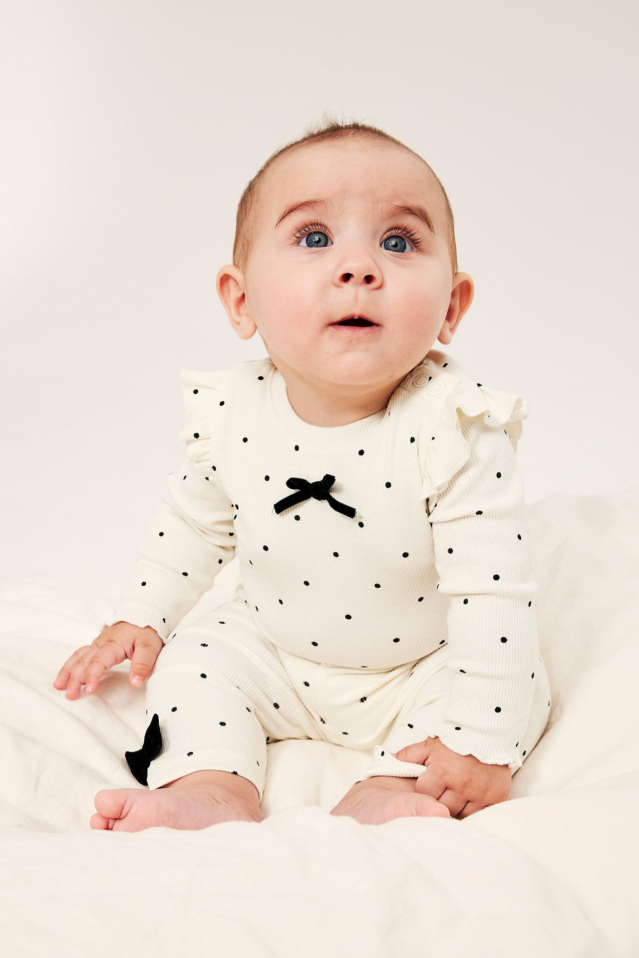 Från framsidan: baby i en off-white ribbad långärmad prickig body med volanger och svart rosett.