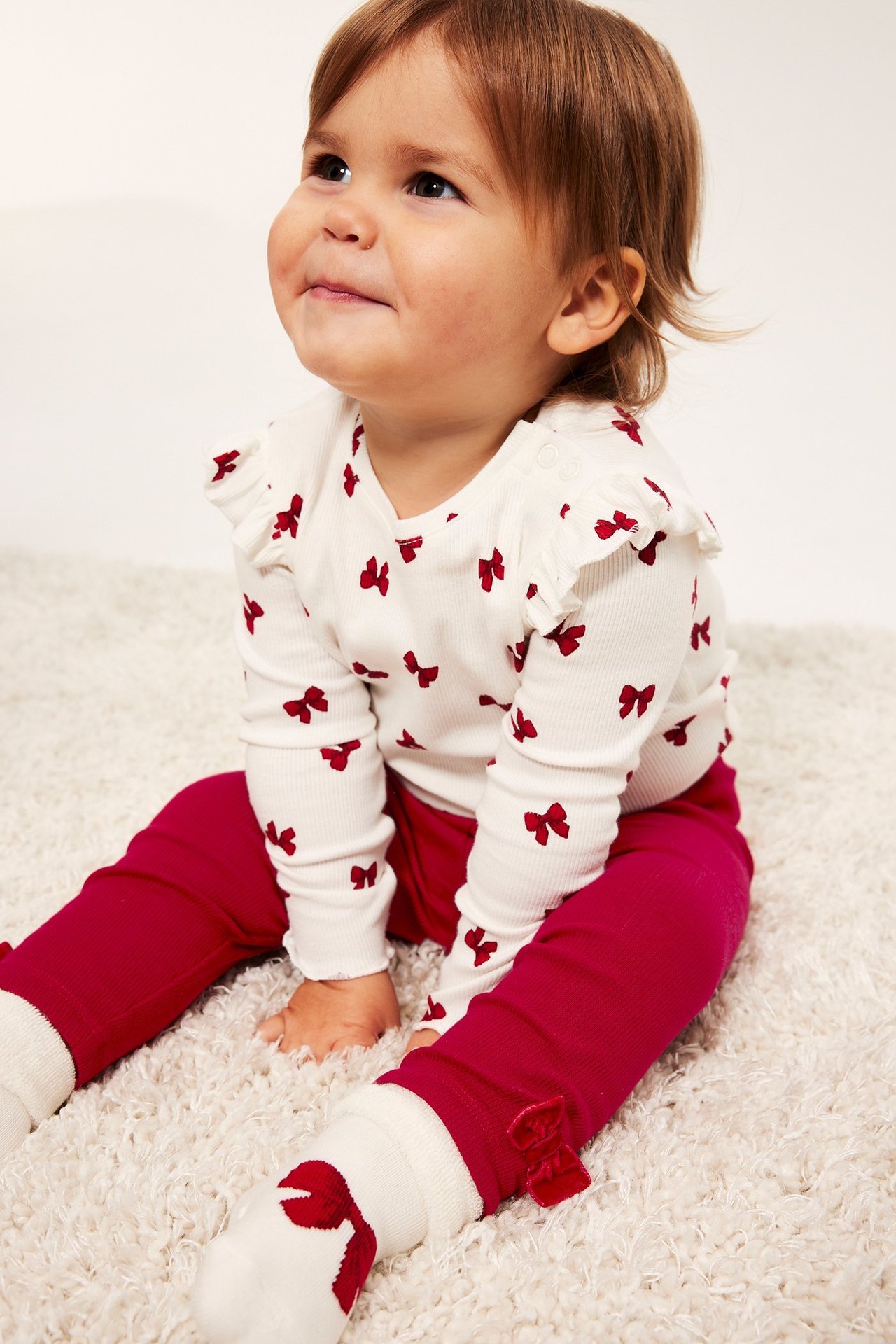 Babyjente i hvit bluse med røde sløyfer og røde leggings, sitter på teppe.