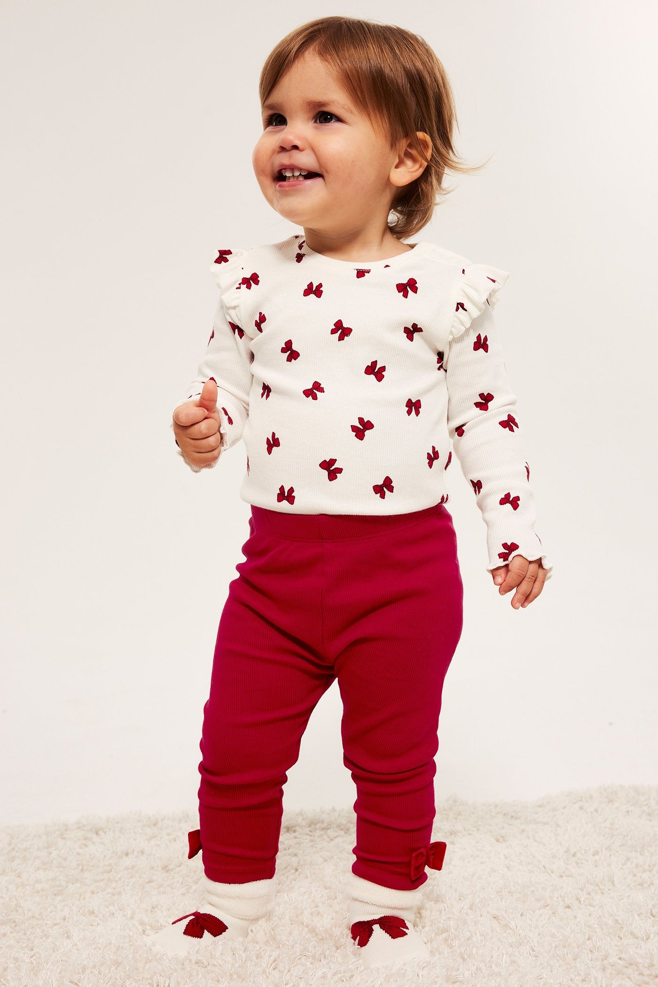 Barn i hvit langermet genser med røde rosetter og røde ribbede leggings.