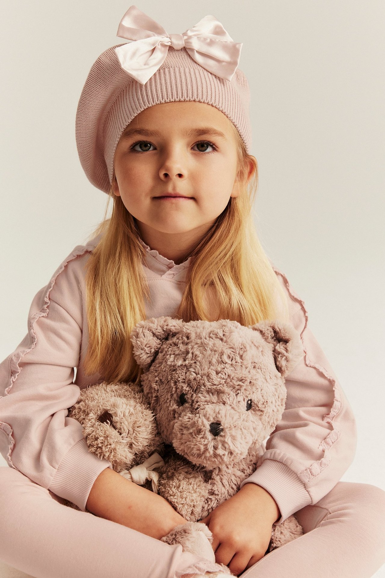 Widok z przodu: Dziewczynka w jasnoróżowej bluzie z kapturem i koronką, pasującym berecie, trzyma misia.