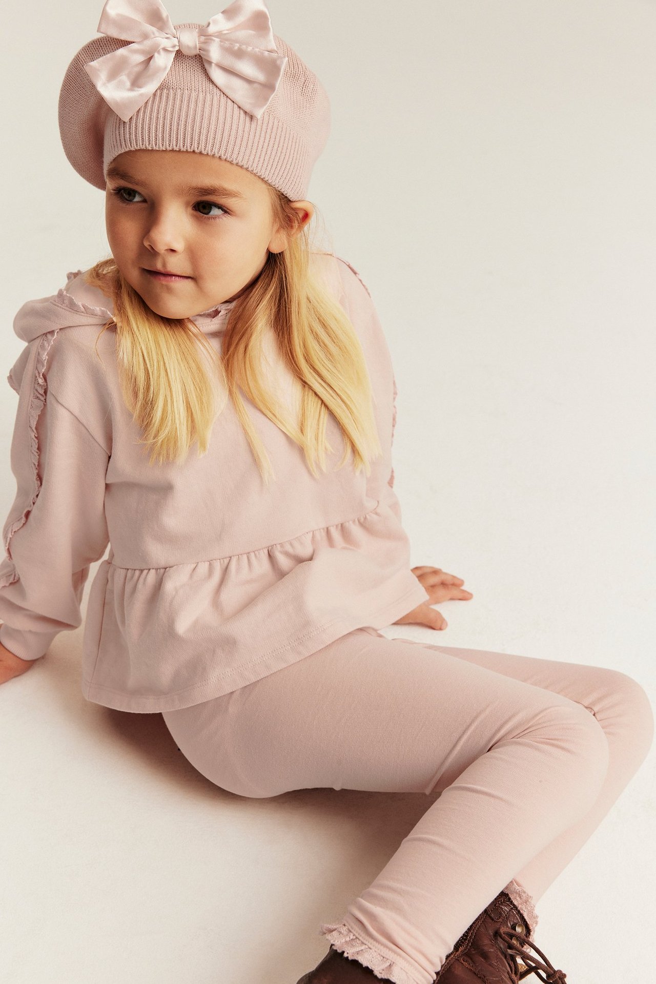 Edestä: Tyttölapsi yllään vaaleanpunainen huppari pitsillä, yhteensopivat leggingsit ja vaaleanpunainen rusettibaretti.