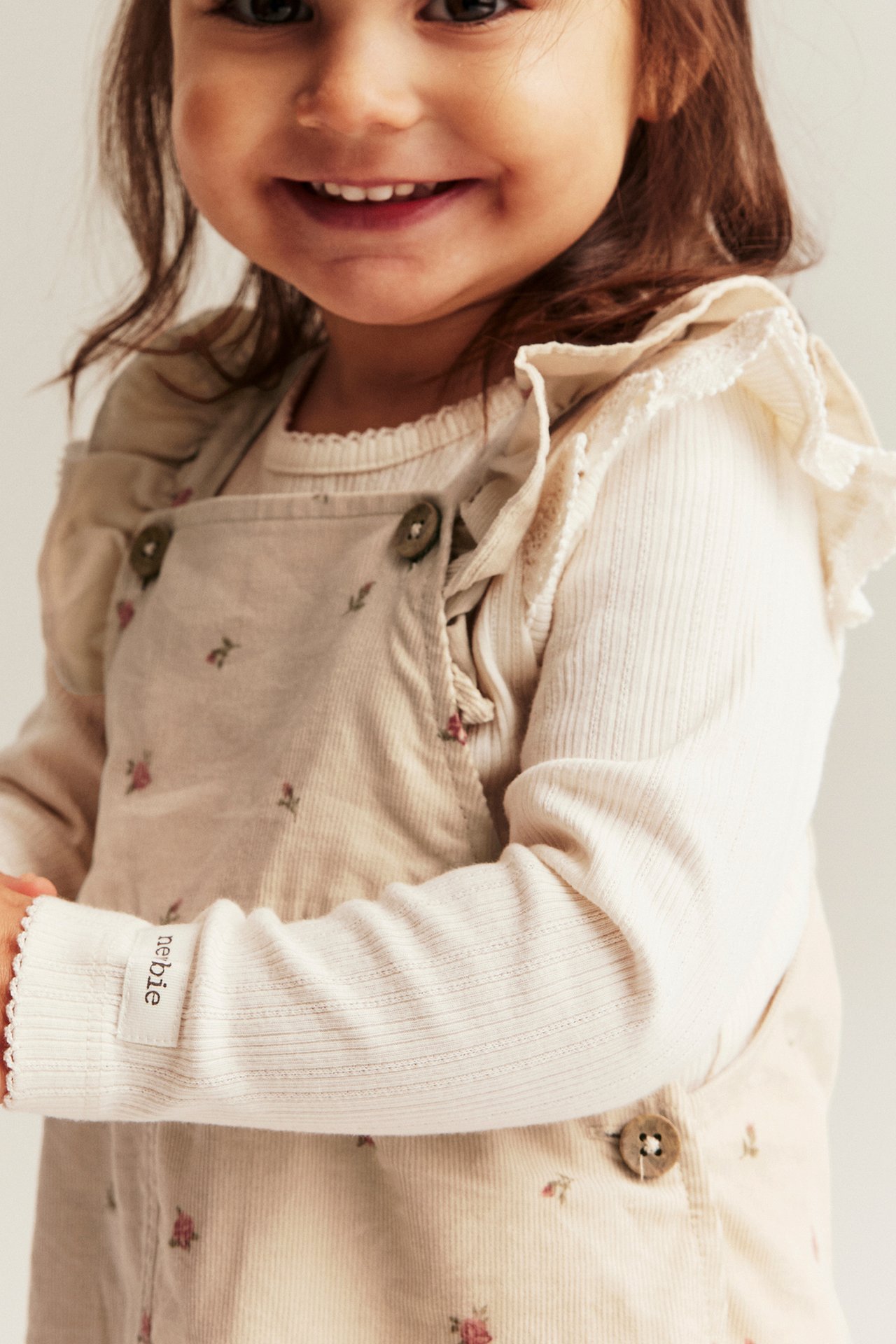 Framifrån, leende barn i beiga blommiga manchesterhängselbyxor över en gräddvit långärmad topp med volanger.