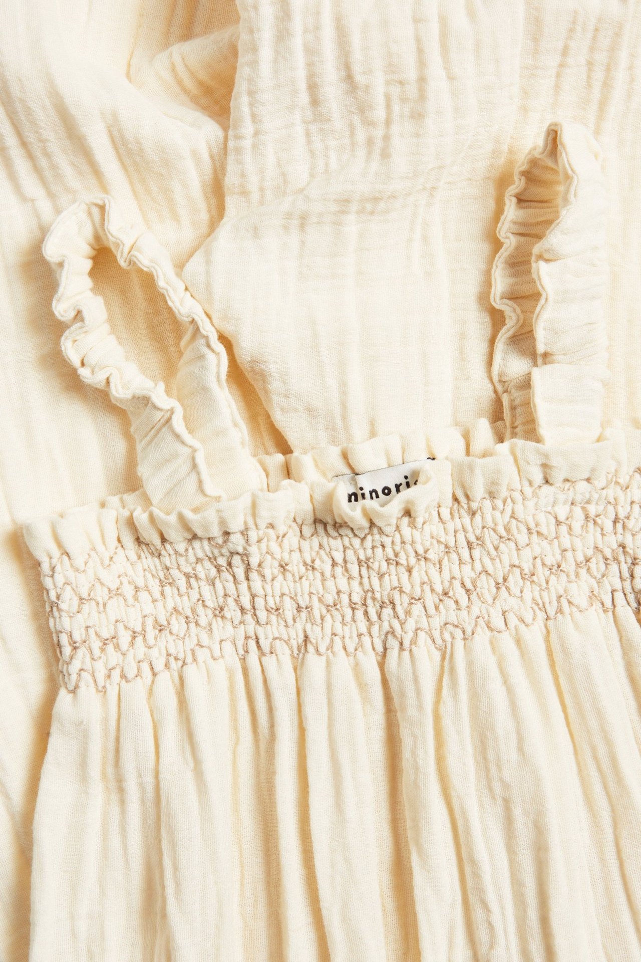 Närbild på beige jumpsuit i bomullskrep med volangaxelband och smockad överdel med brun söm.