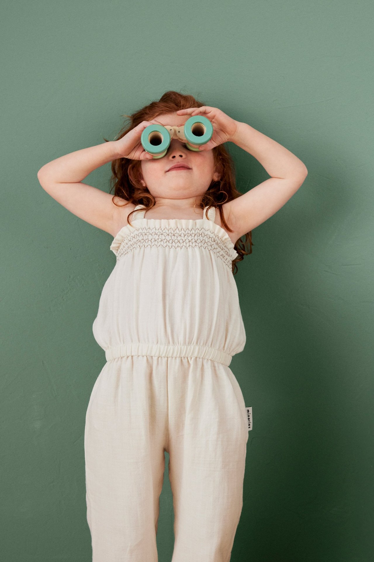 Framifrån: ett barn i beige bomullscrepe-jumpsuit med smockad topp, håller gröna kikare.