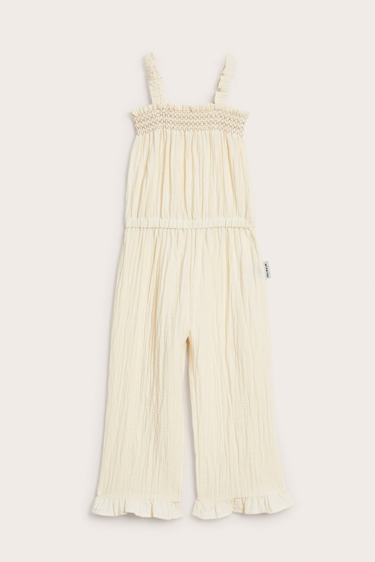 Framsida av en beige bomullskrepp-jumpsuit för barn med smockad topp, resår i midjan och volangfållar.