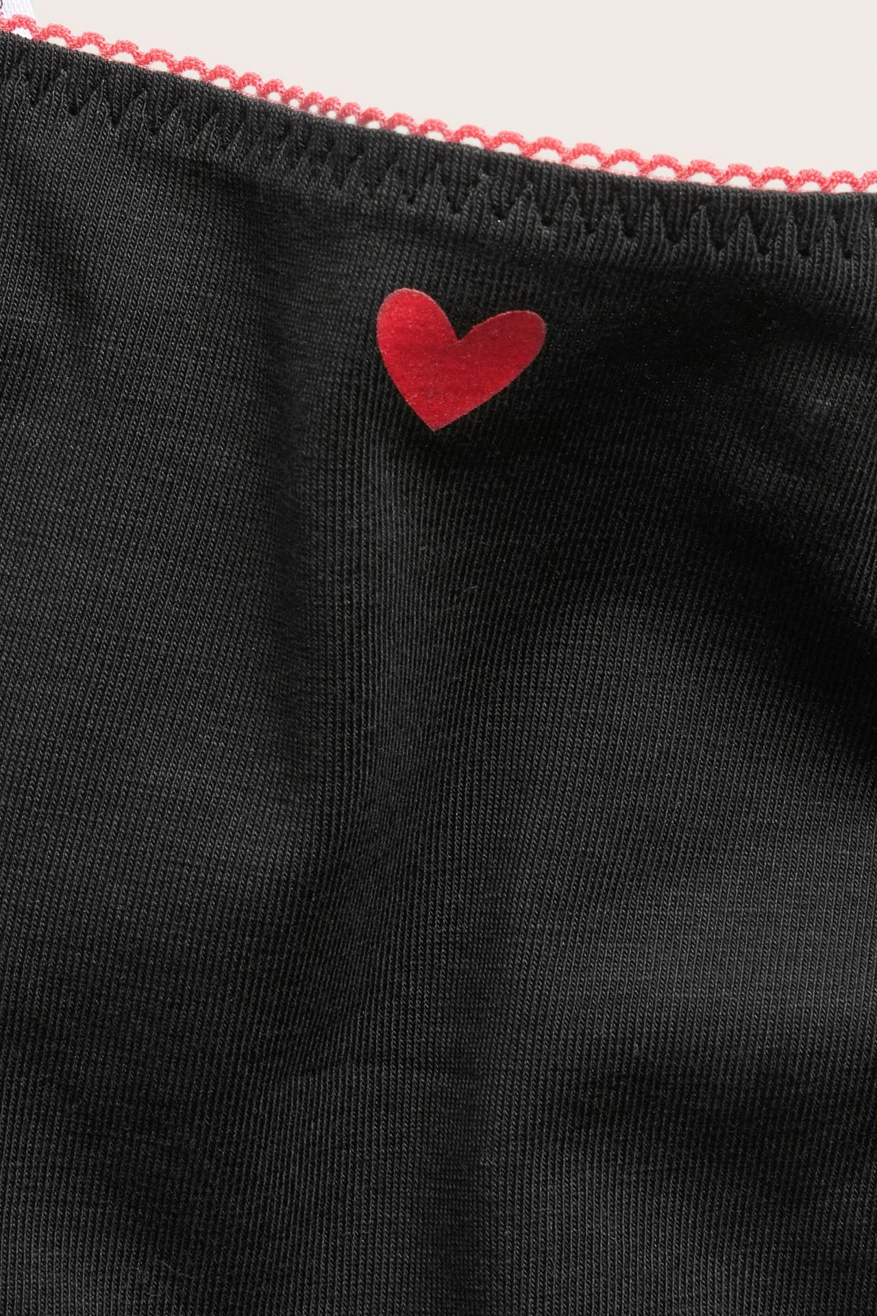 Nærbilde av en svart stringtruse i bomullsblanding med rødt hjerte og rosa picot-elastikkant.