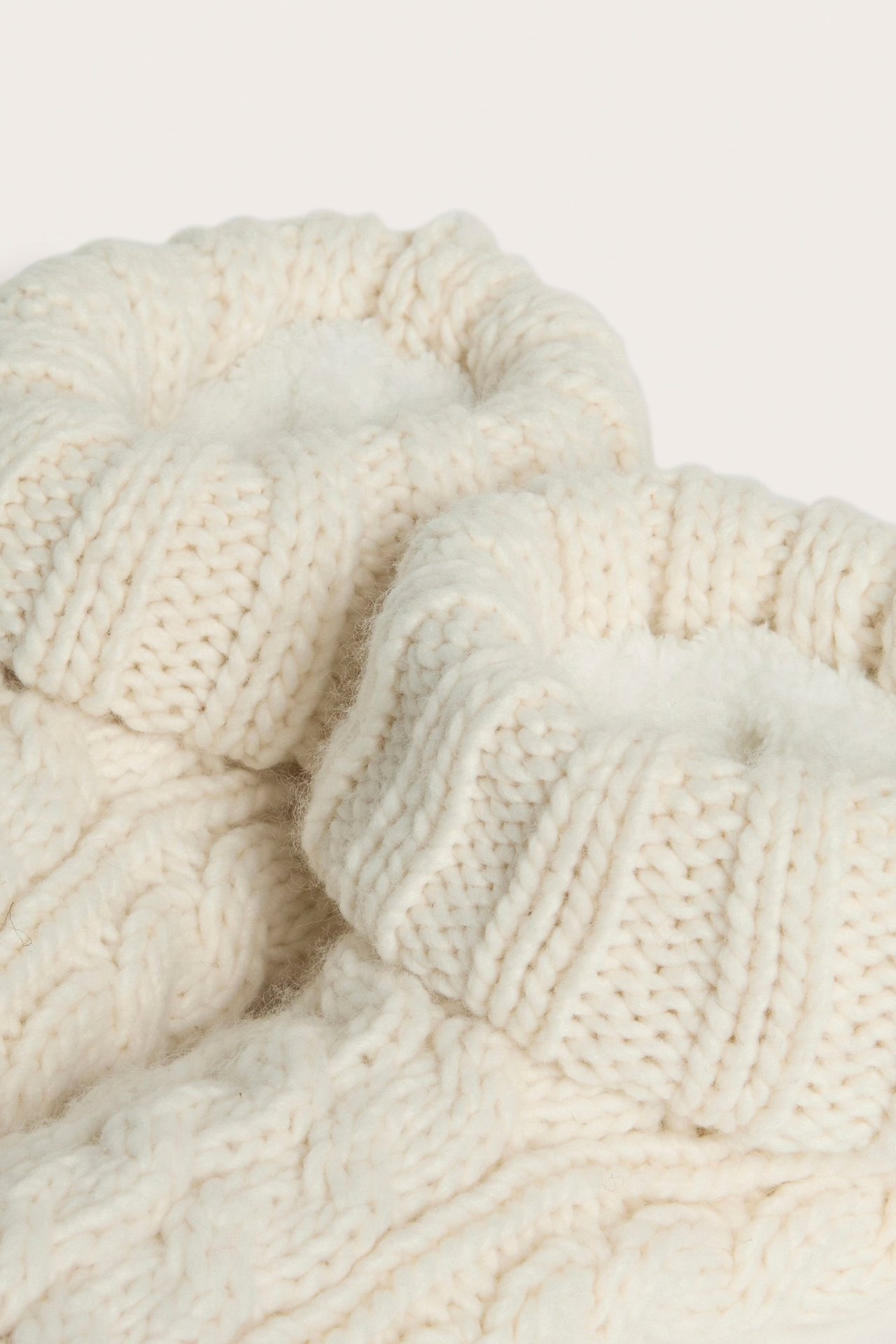 Zbliżenie na dwie jasnobeżowe kapcie ze splotem warkoczowym, z miękkim polarem i wywiniętymi mankietami.