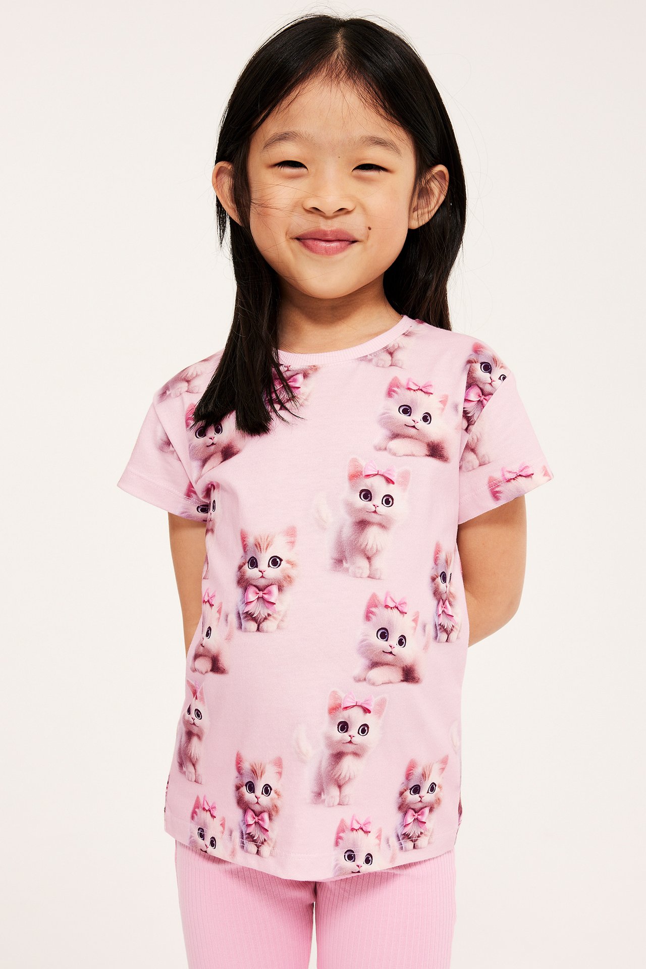 Widok z przodu dziecka w różowym bawełnianym T-shircie z nadrukiem kotków i różowych legginsach w prążki.