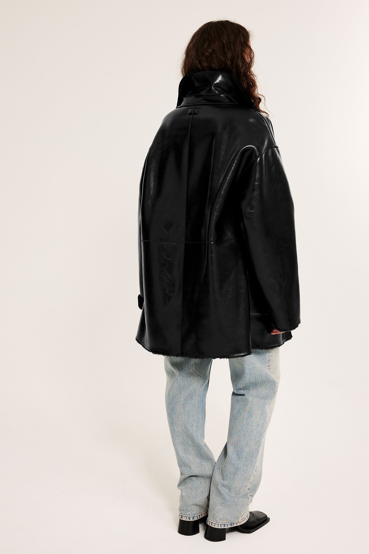 Ryggvy av kvinna i svart överdimensionerad jacka i skinnimitation med fårpäls, ljusa jeans och svarta skor.