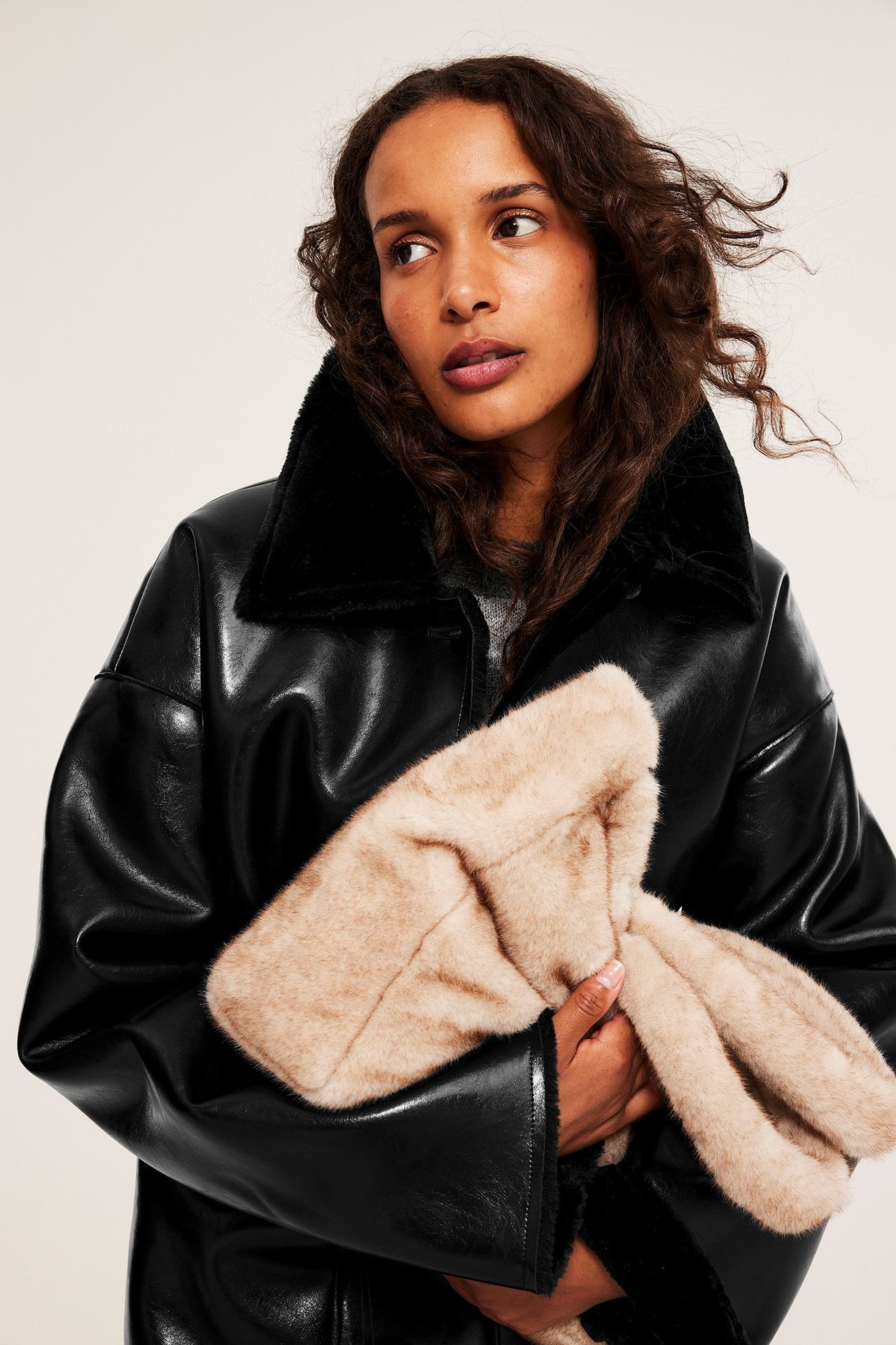 Lähikuva naisesta mustassa keinonahkatakissa tekoturkiksella, pitäen vaaleanbeigettä tekoturkislaukkua.