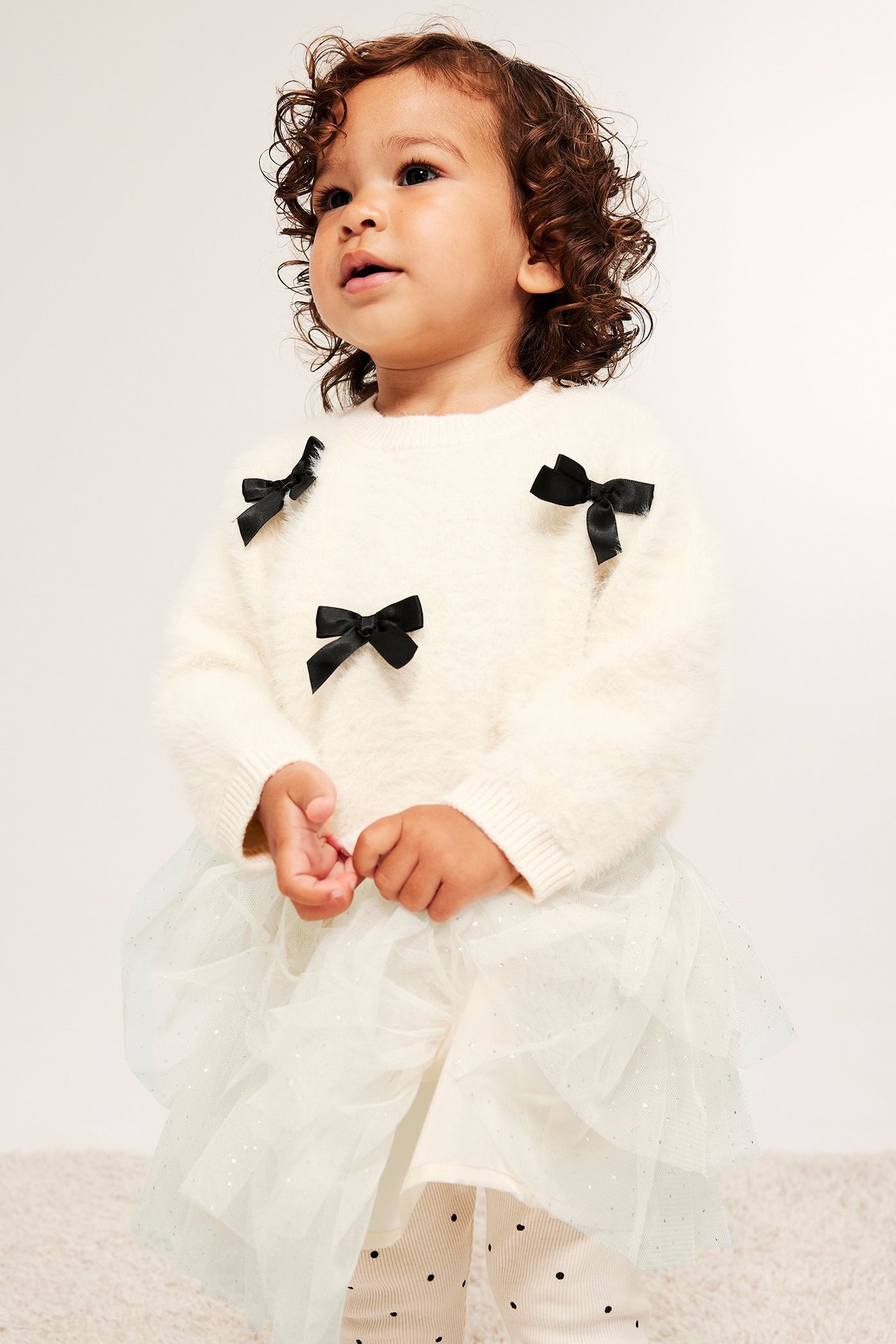 Forfra: baby i et lys beige glitrende mesh-skjørt, en kremfarget fluffy genser med sorte sløyfer og prikkete leggings.