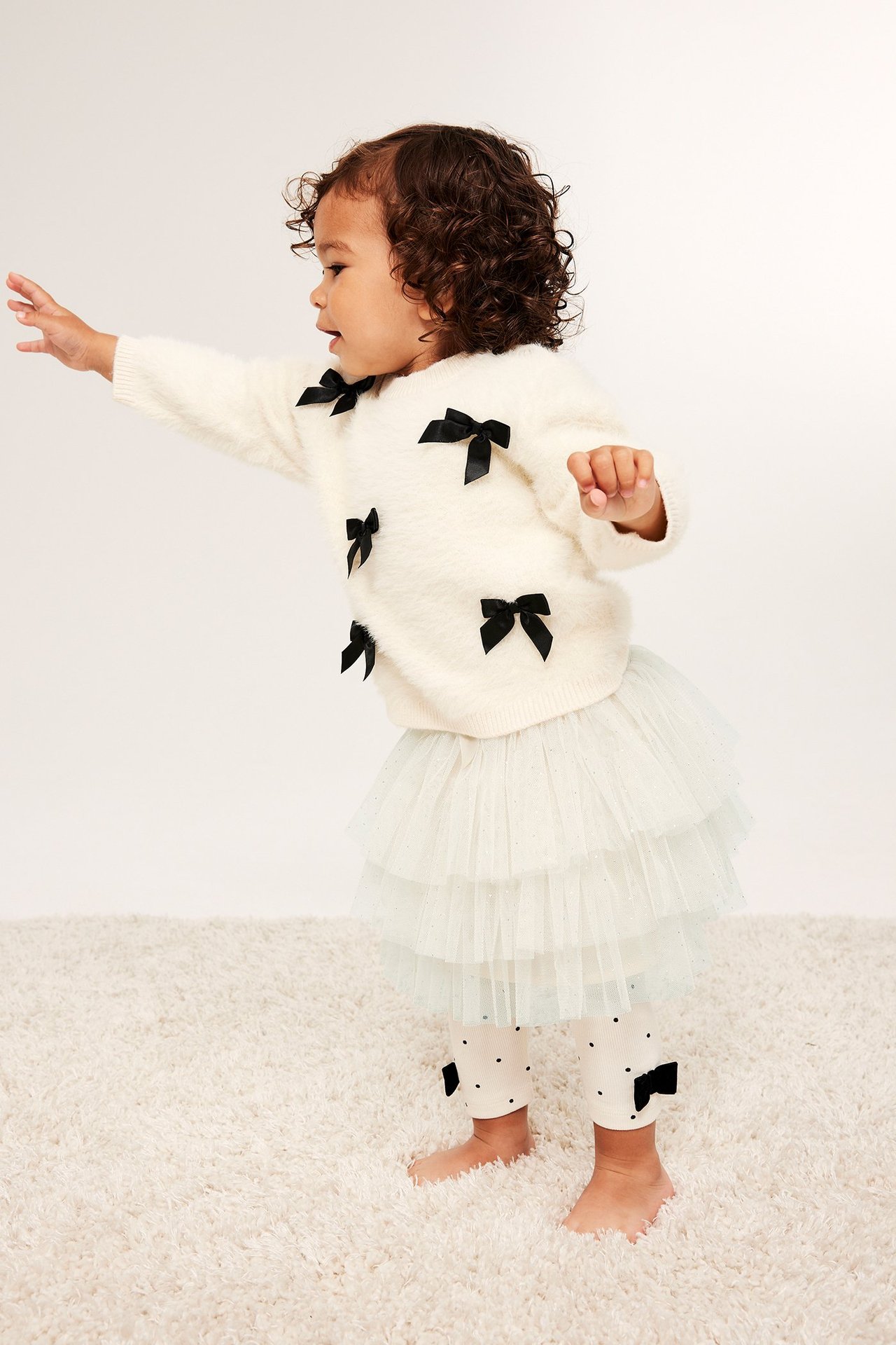 Sidebilde av baby: lys beige glitter meshskjørt, kremgenser med svarte sløyfer og hvite prikkete leggings.