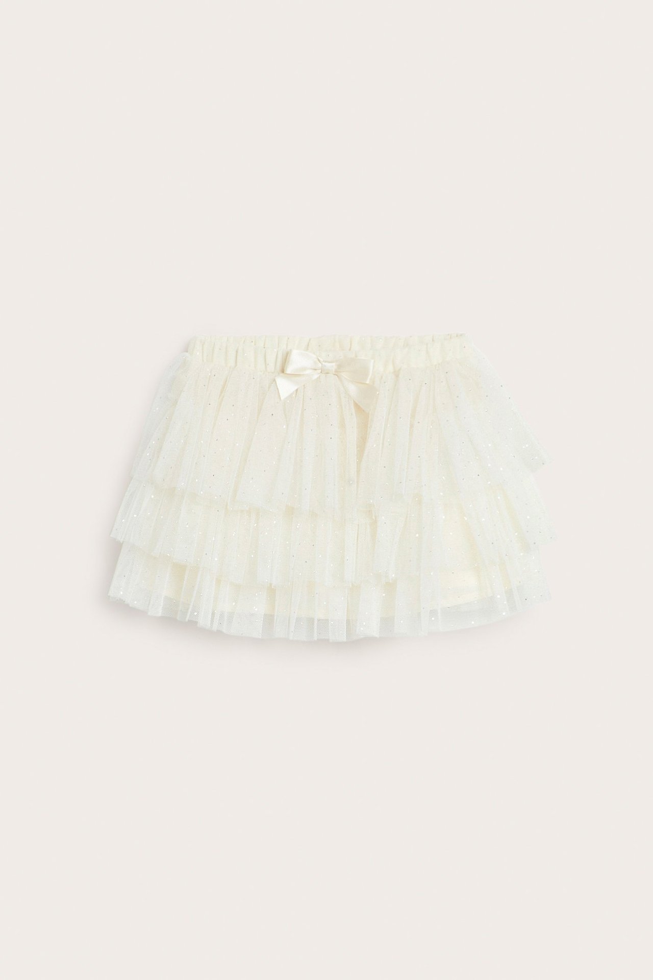 Frontvisning av et babys lys beige, lagdelt meshskjørt med glittertrykk og en liten sløyfe.