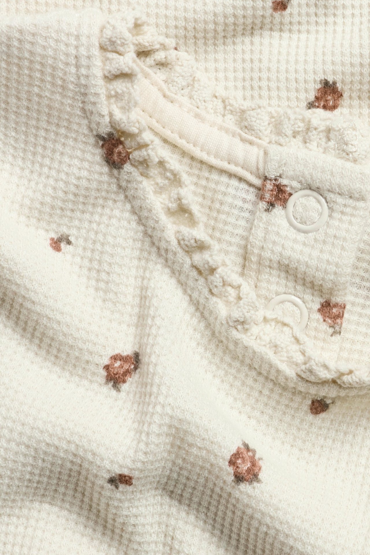 Zbliżenie na jasnobeżowy top niemowlęcy z dzianiny waflowej w kwiatowy wzór, z koronką i zatrzaskami.