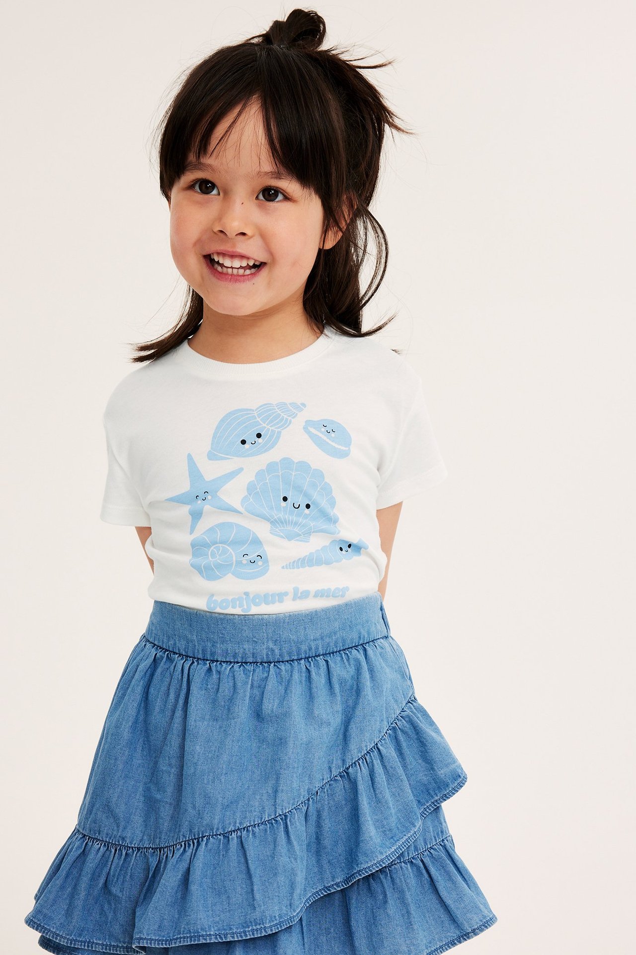 Dziecko w białym T-shircie z niebieskim nadrukiem muszelek i niebieskiej jeansowej spódnicy z falbanami.