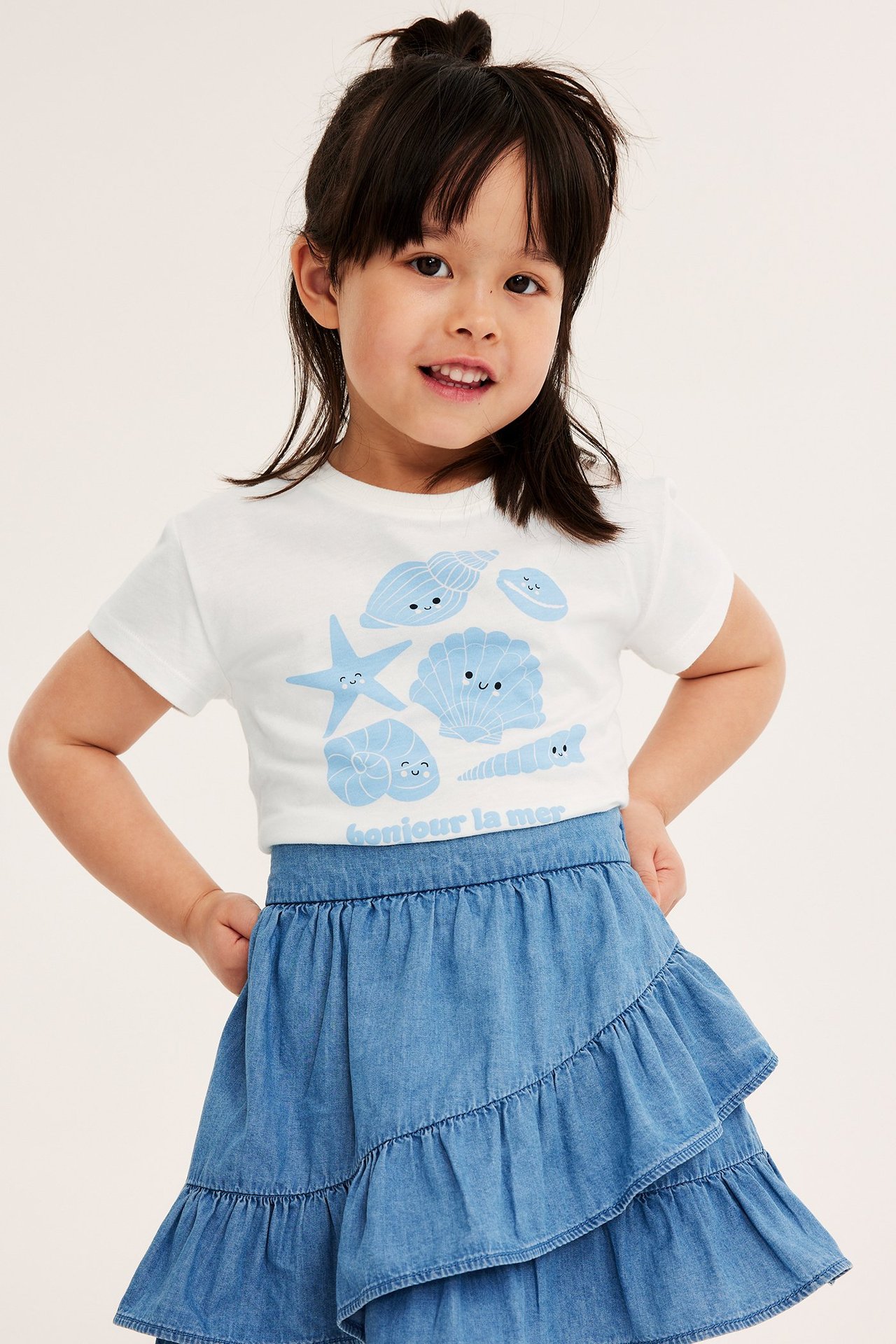 Dziecko w białym T-shircie z niebieskim nadrukiem muszelek i niebieskiej spódnicy z falbanami.