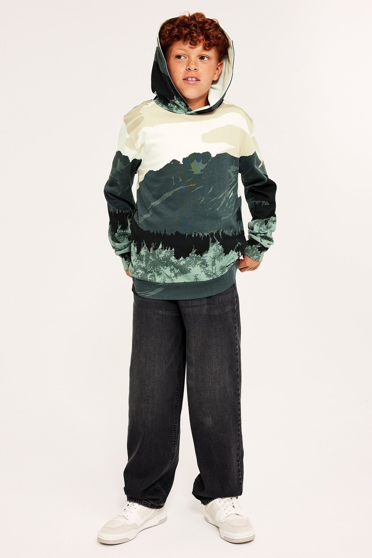 Widok z przodu: Dziecko w beżowo-zielono-czarnej bluzie z kapturem z nadrukiem gór, ciemnoszarych dżinsach i białych trampkach.