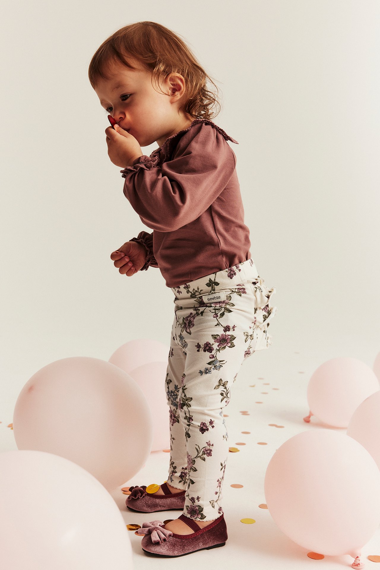 Sidebilde av et barn i lys beige blomstrete leggings med volanger bak, en rosa topp og fløyelssko.