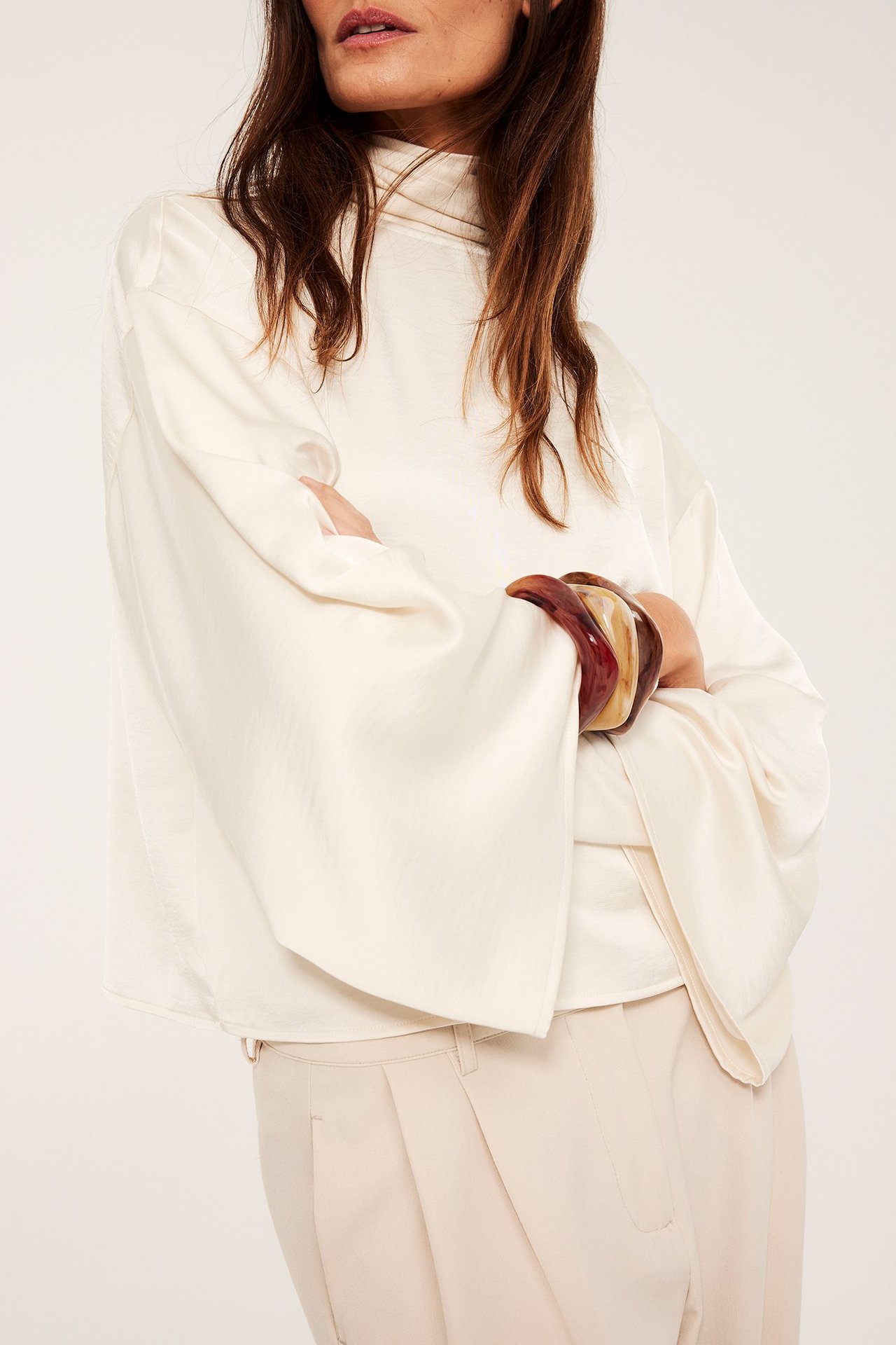 Lähikuva edestä: Nainen, kolme ruskeaa marmoroitua muoviranneketta, luonnonvalkoinen paitapusero, vaaleanbeiget housut.