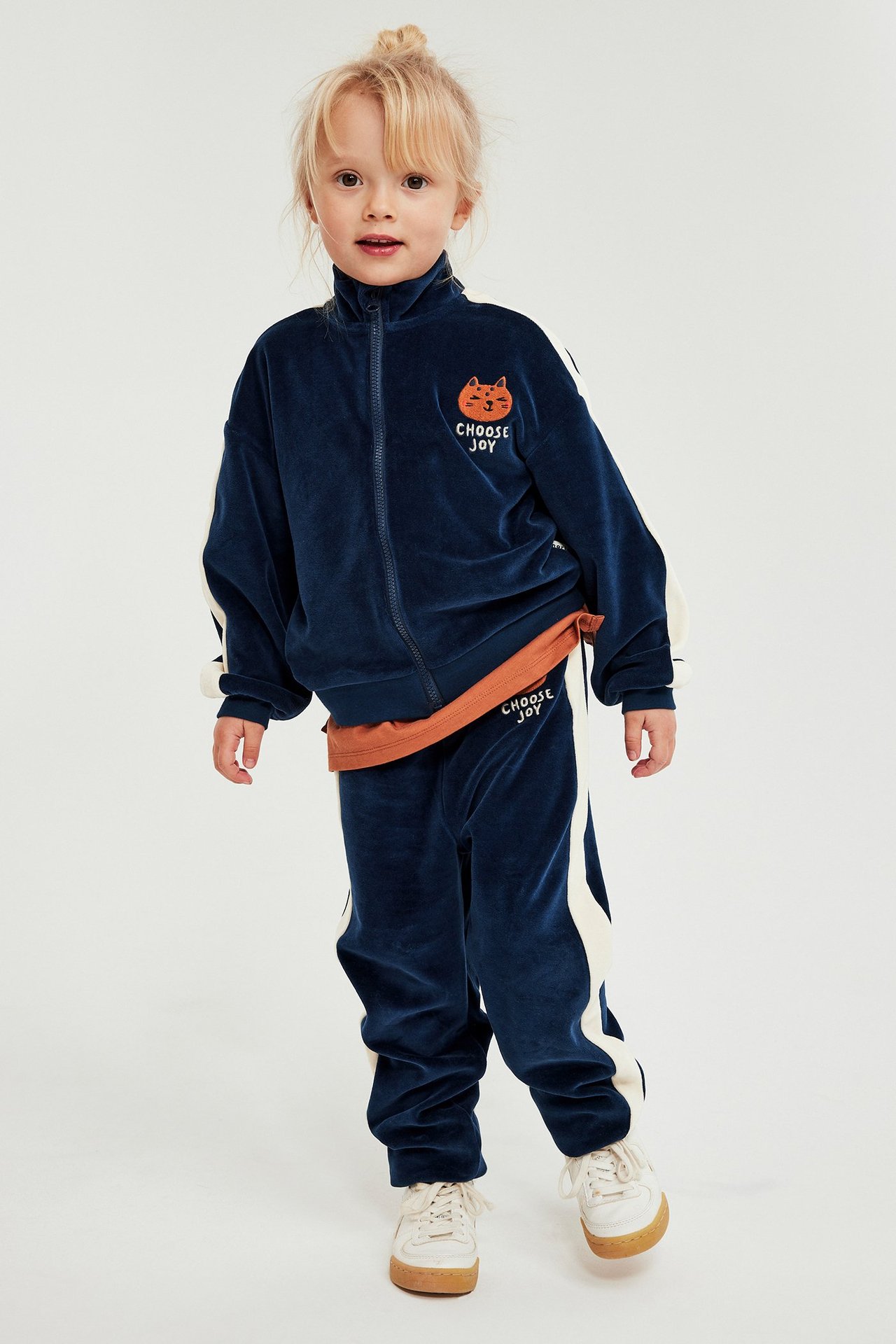 Kid bär mörkblå velourtröja med dragkedja, vita revärer, brodyr, matchande byxor och orange t-shirt.