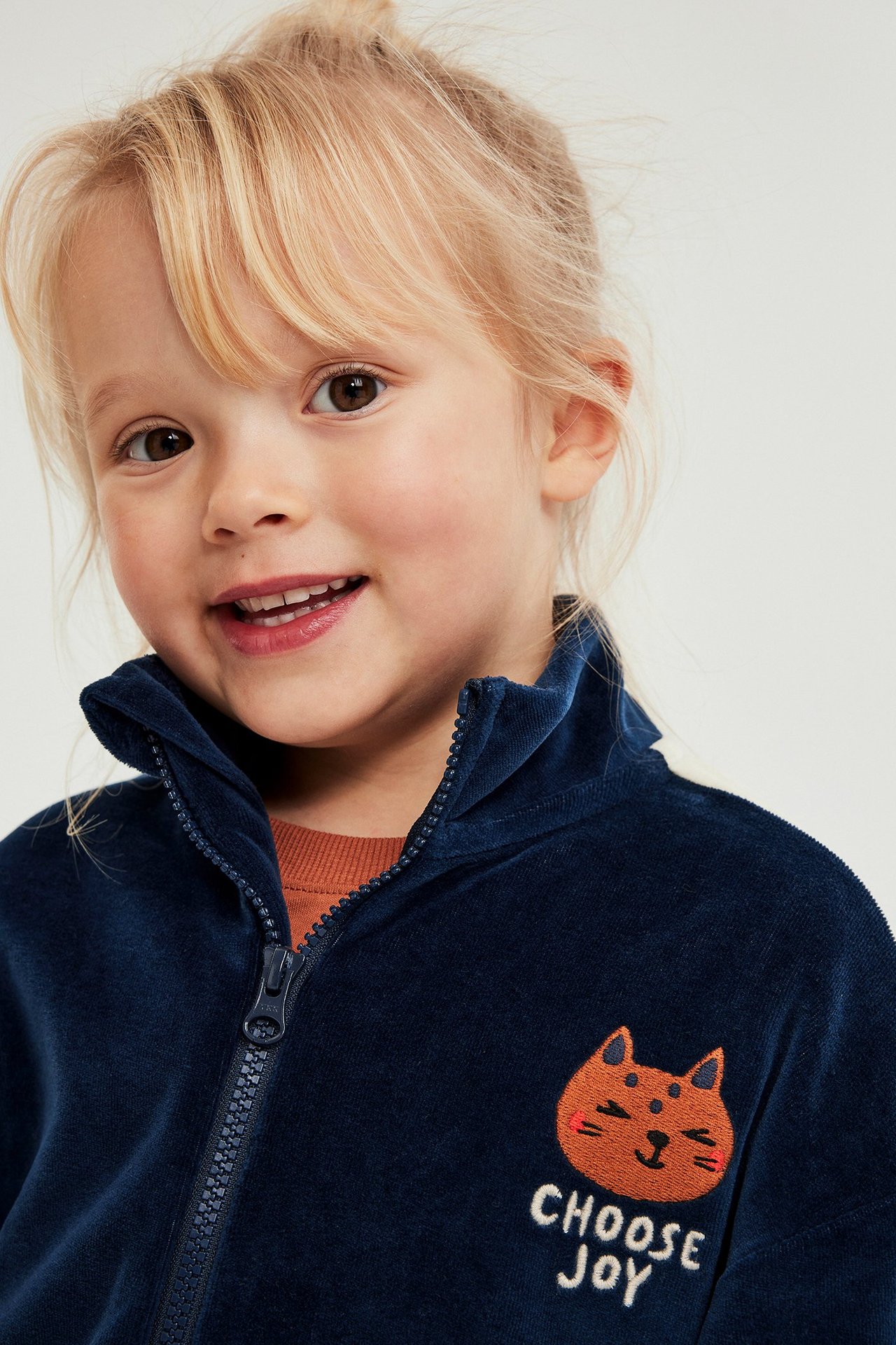 Närbild på ett barn i en mörkblå velourtröja med dragkedja och kattbrodyr, över en orange tröja.