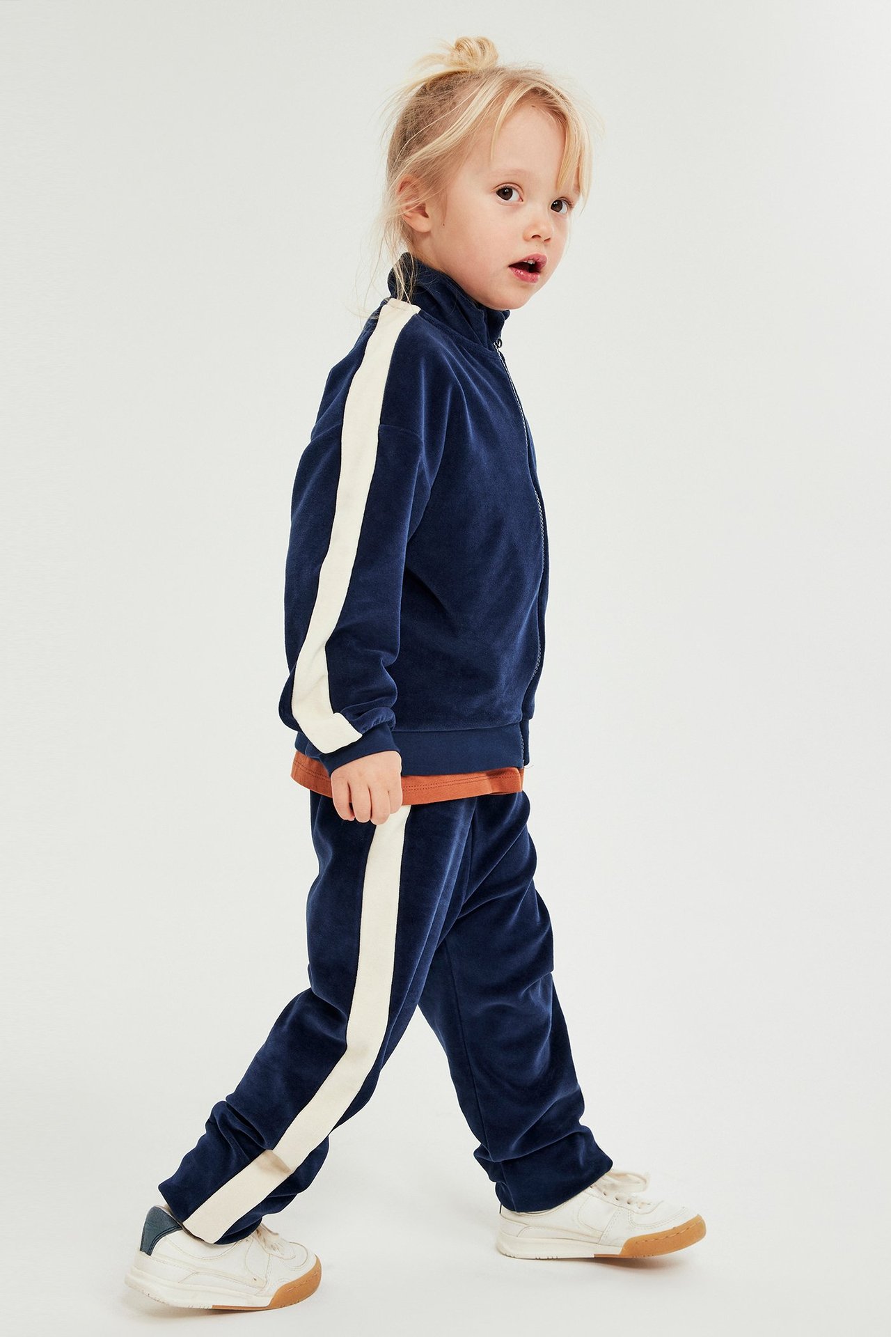 Sivukuva lapsesta tummansinisessä veluuriverryttelyasussa, kermanväriset sivuraidat ja valkoiset tennarit.