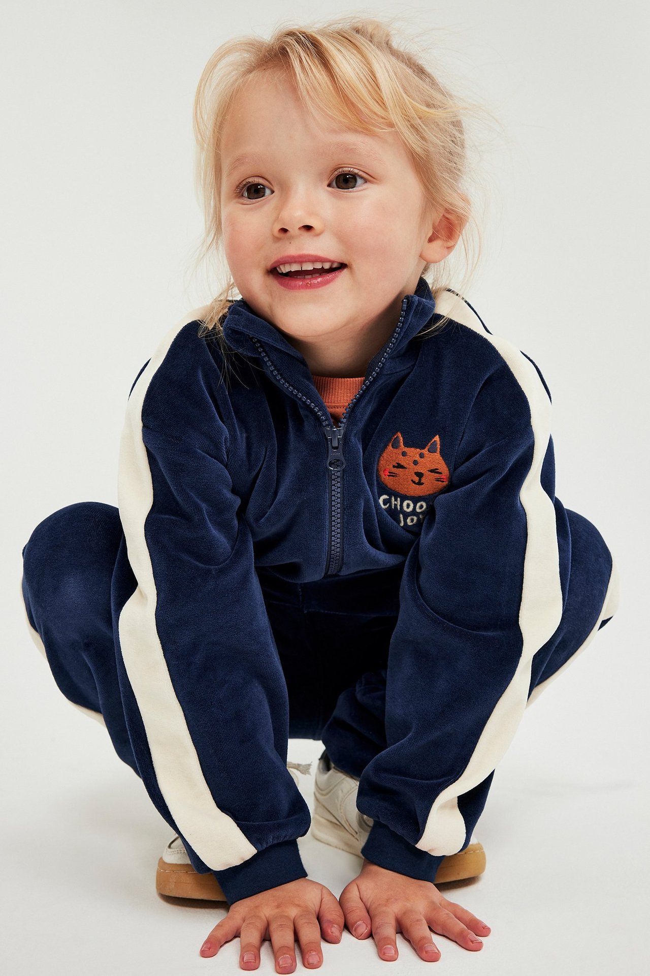 Edestäpäin: lapsi tummansinisessä veluuriverryttelyasussa, jossa vaaleat sivuraidat ja kissakirjonta.