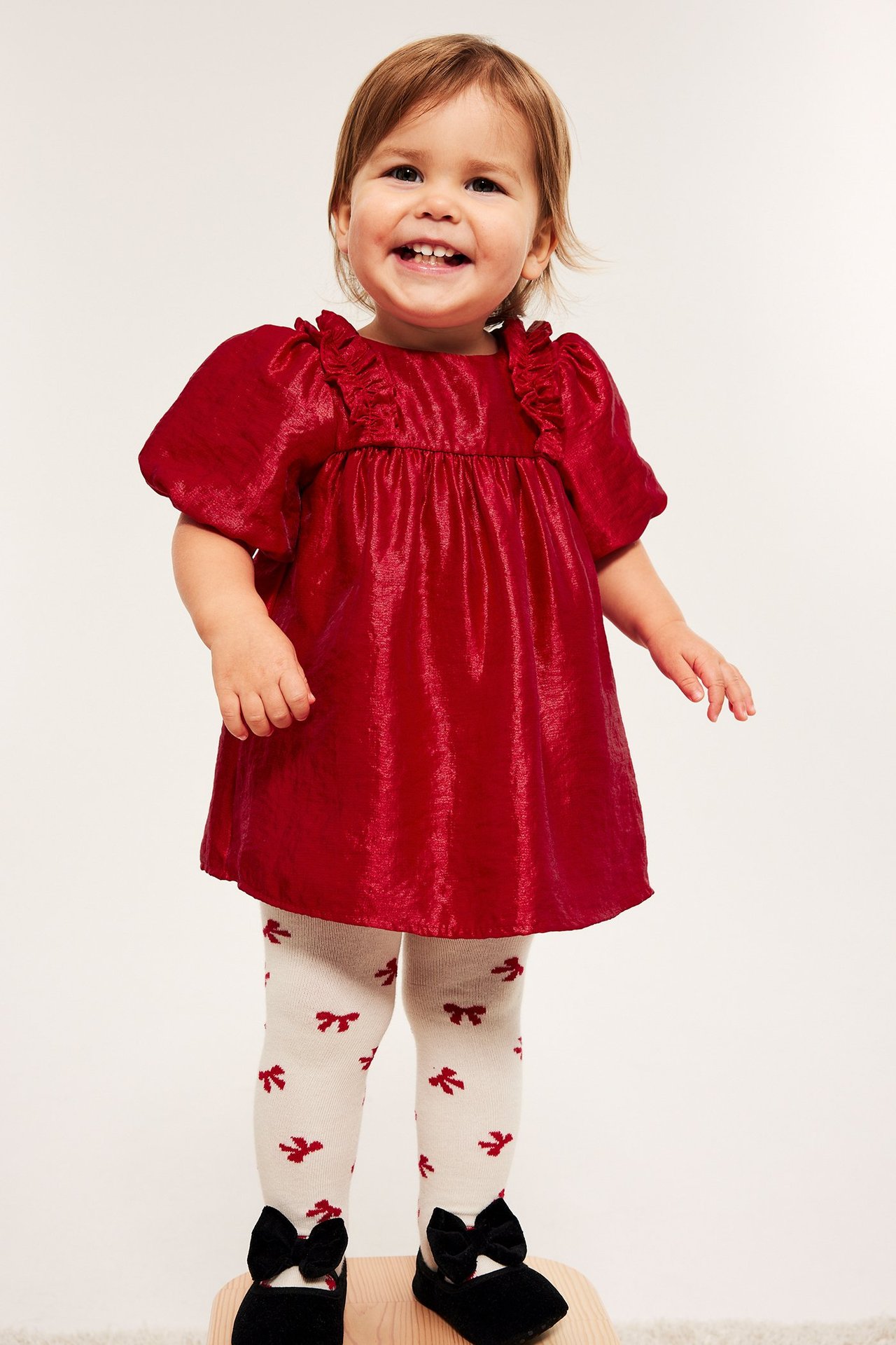 Jente i rød viskosekjole med puffede ermer og volanger, knapper i nakken, smiler til kamera.