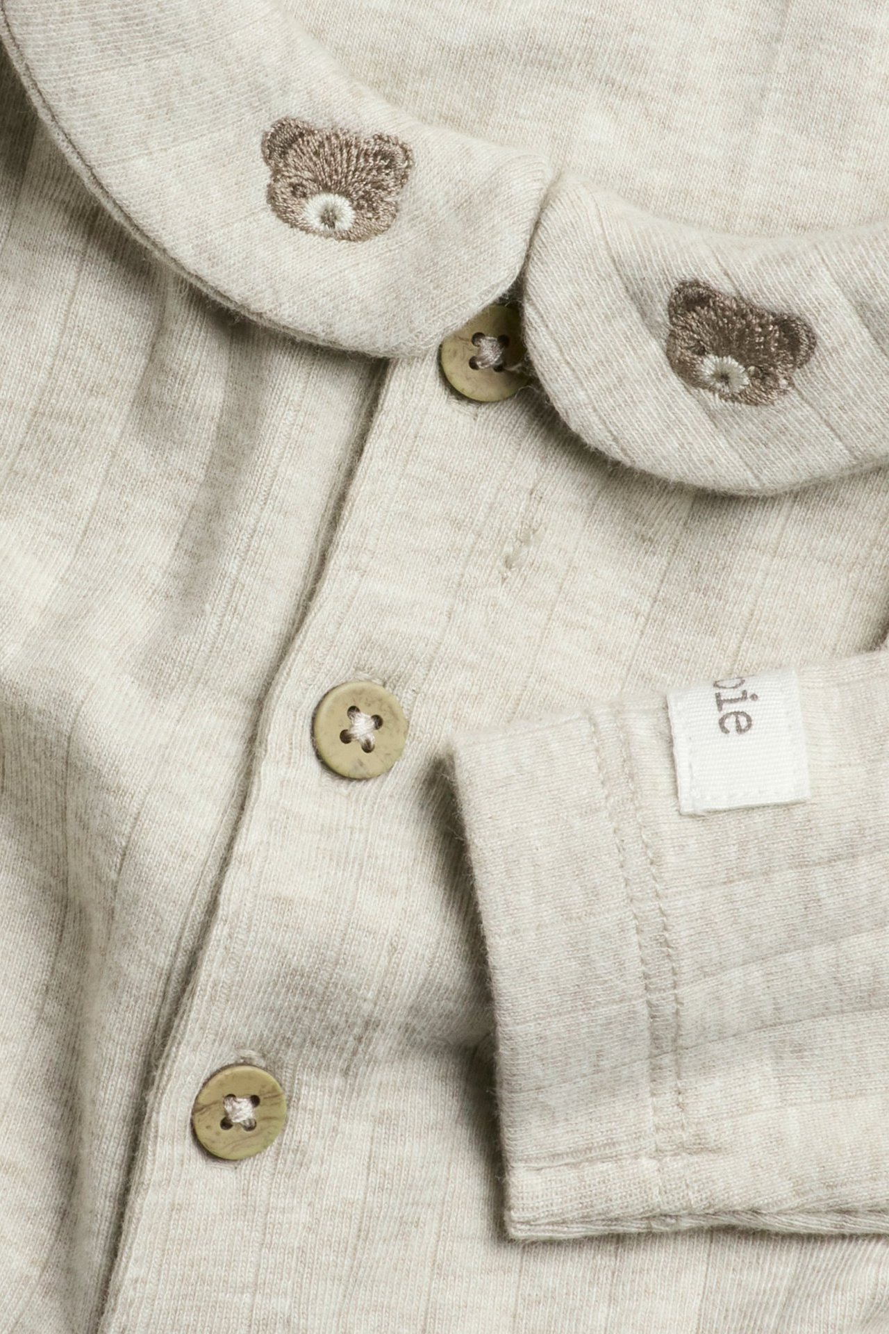 Nærbilde av en beige ribbet babybody med krage, teddybjørn-broderi og knapper foran.
