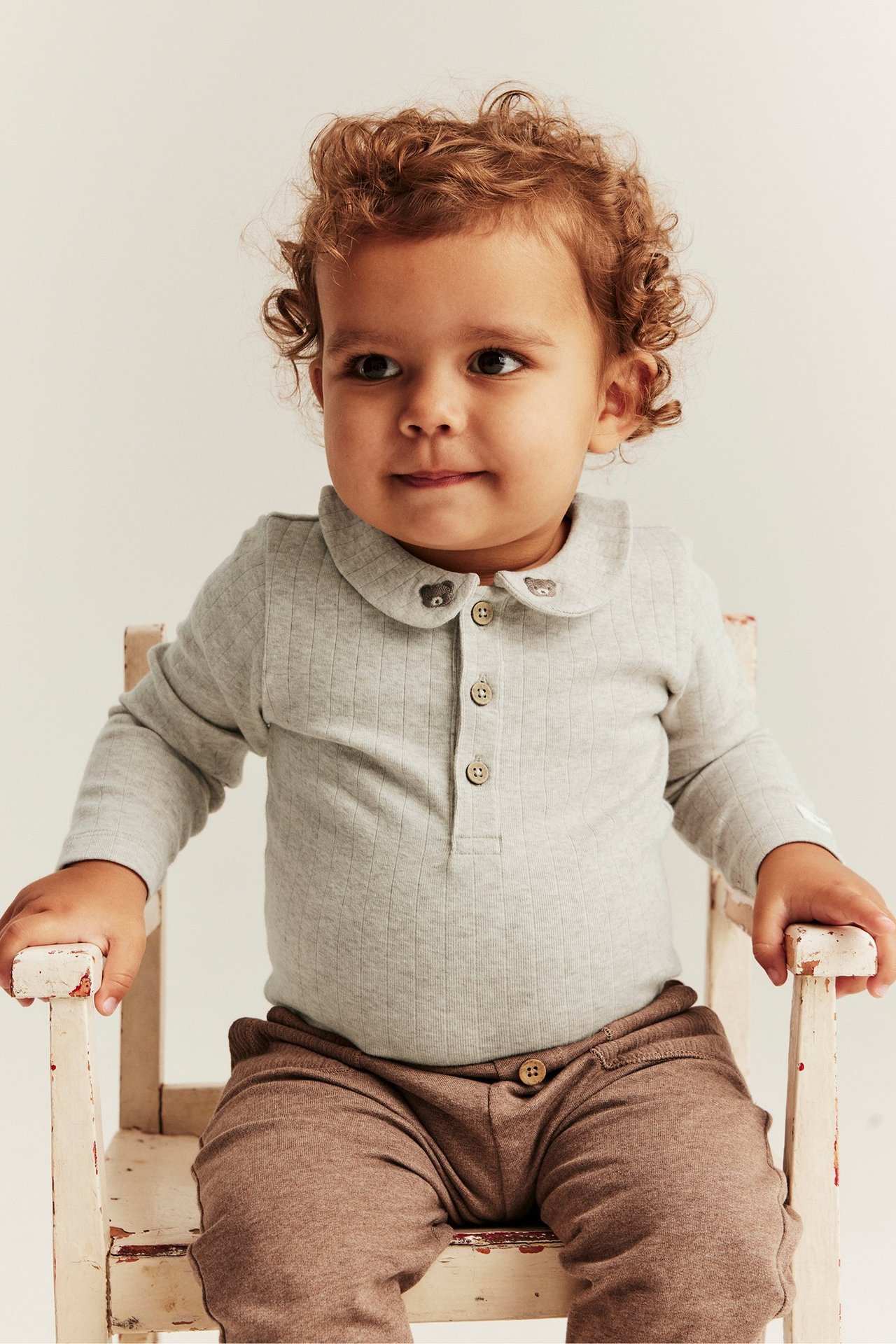 Forfra: baby i lys grå ribbet body med teddybjørn-brodert krage og brune bukser.