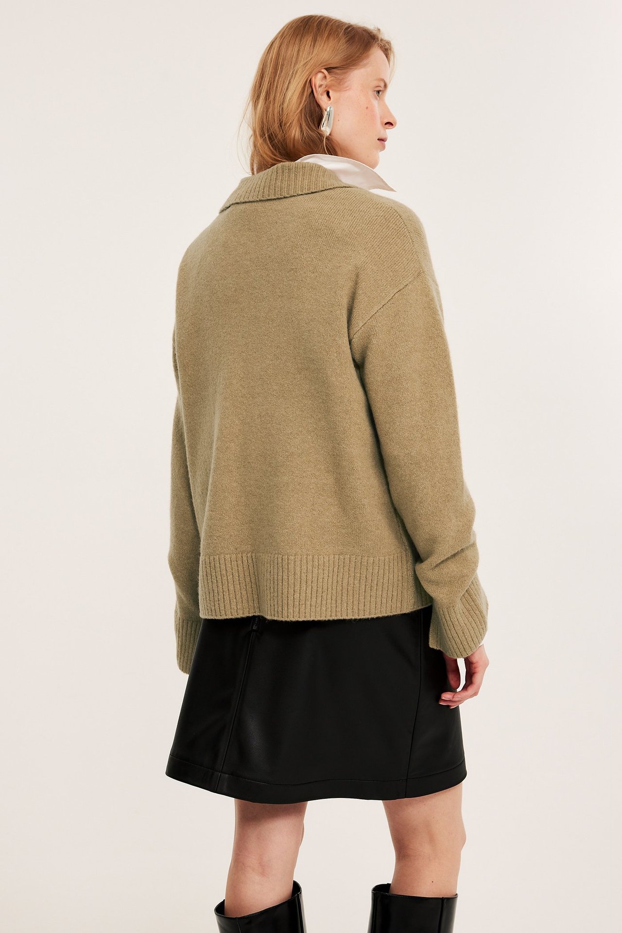 Bakfra, kvinne i melert beige strikket pologenser over hvit skjorte, svart skjørt og støvler.