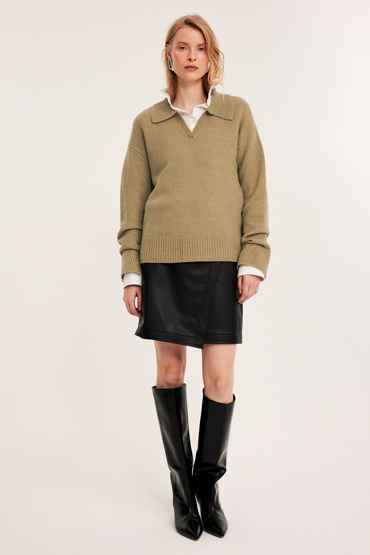 Front view: Kvinna i beige melerad stickad tröja, vit skjorta, svart skinnkjol, svarta stövlar.