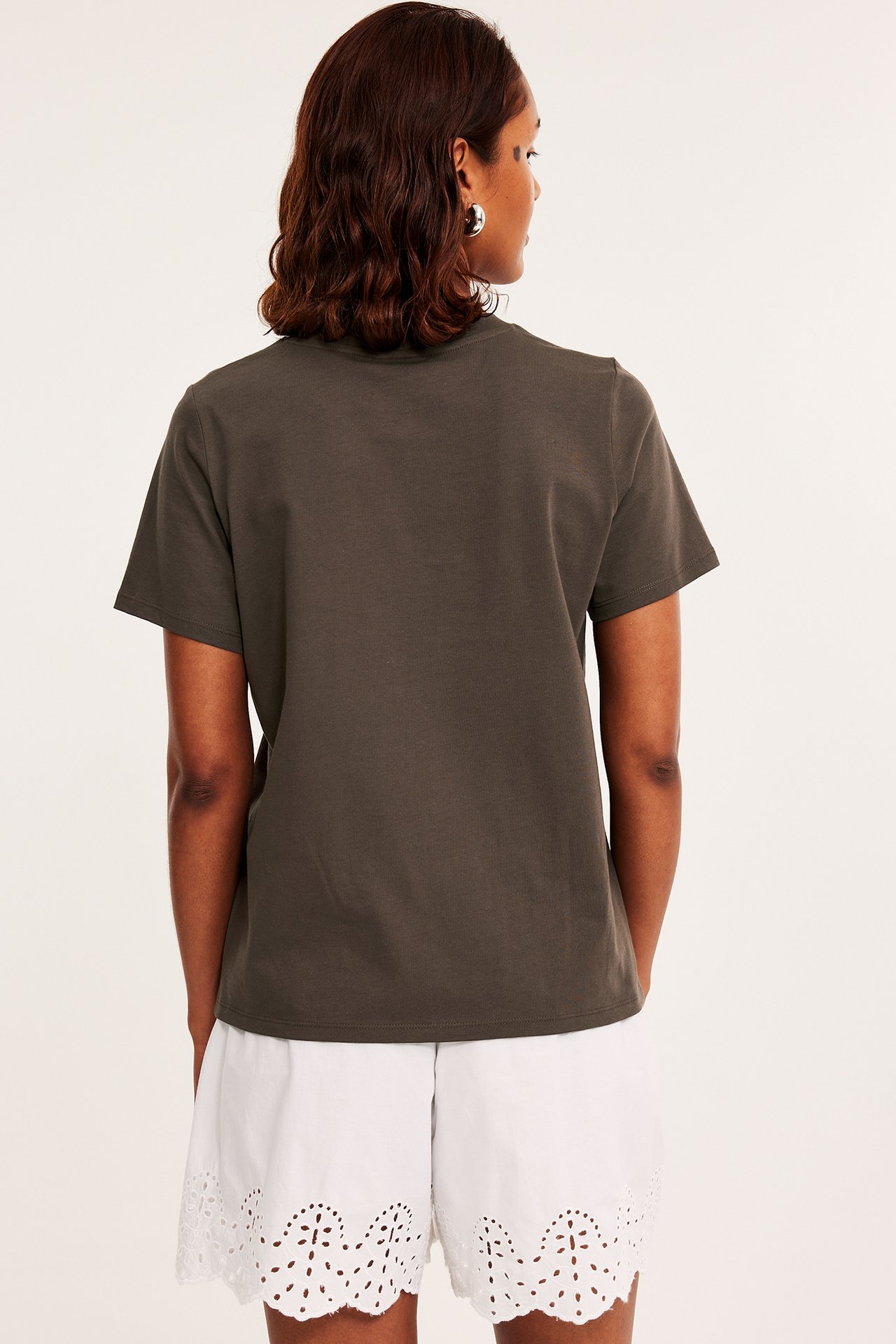 Bakifrån, kvinna i mörkbrun, rakt skuren bomulls-T-shirt och vita shorts med broderade hål.