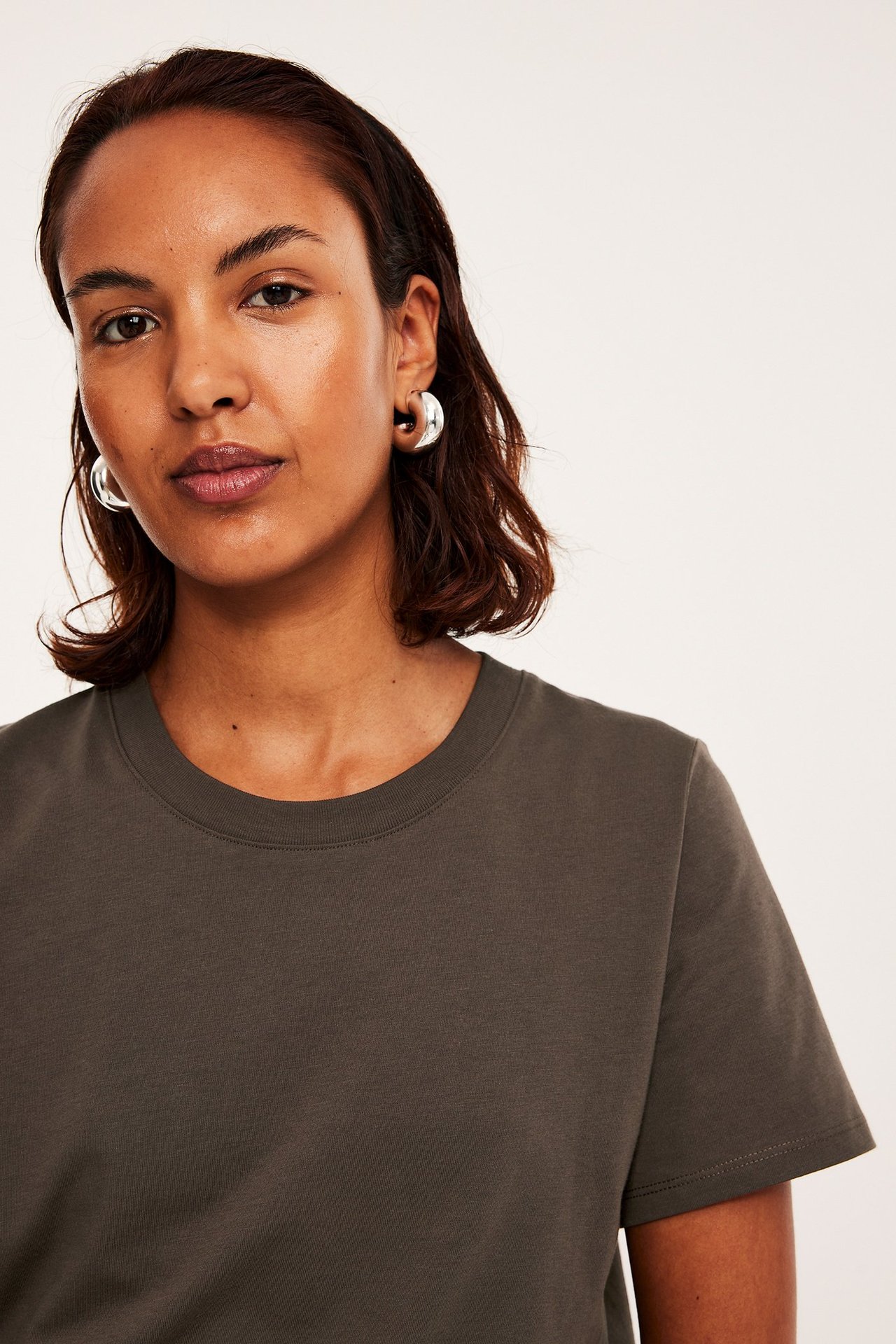 Nærbilde forfra av en kvinne i mørkebrun, rett passform bomulls-T-skjorte og sølvfargede ringøreringer.