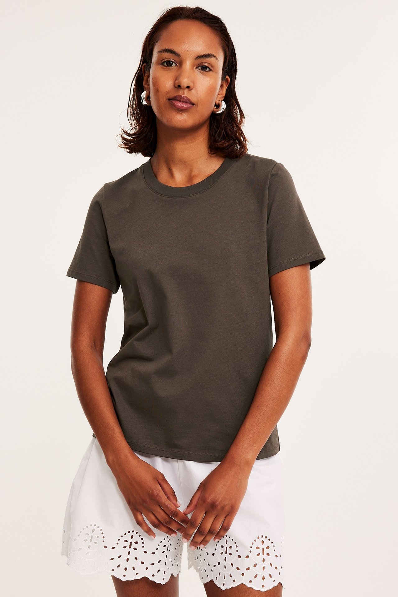 Forfra, kvinne i mørkebrun bomulls-T-skjorte, hvite broderte shorts og sølvfargede ringøreringer.