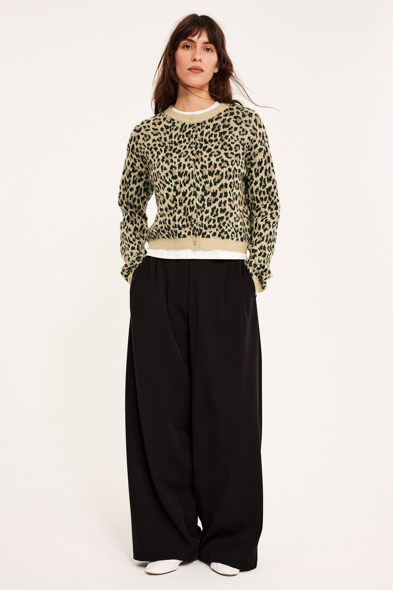Edestäpäin: nainen beigessä leopardikuvioisessa neuletakissa, valkoisessa t-paidassa, mustissa leveissä housuissa ja valkoisissa kengissä.