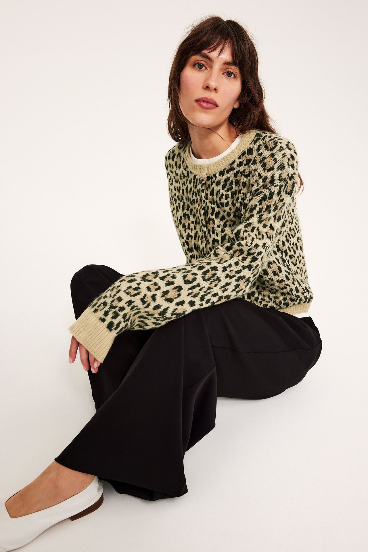 Edestä: Nainen beigessä leopardineuletakissa, valkoisessa topissa, mustissa leveissä housuissa ja valkoisissa kengissä.