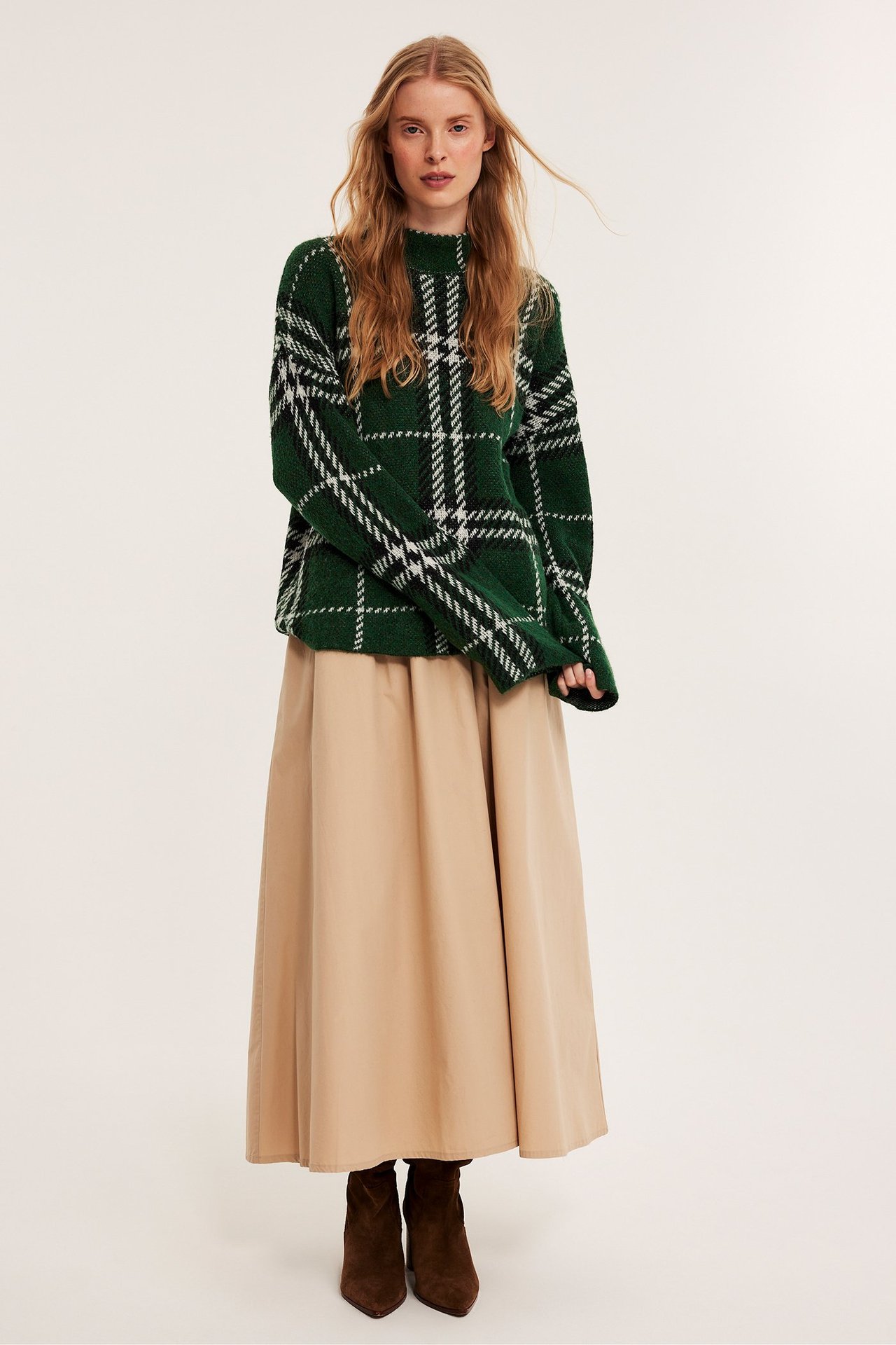 Framifrån: kvinna i mörkgrön rutig stickad pullover, lång beige kjol och mörkbruna ankelstövlar.