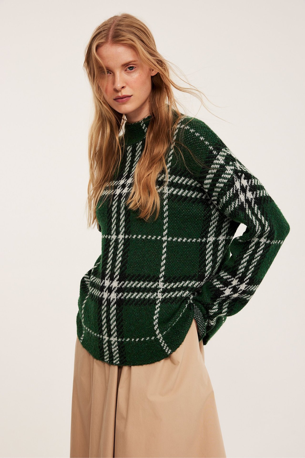 Edestä: Nainen yllään tummanvihreä ruudullinen neulepusero ja vaaleanbeiget leveälahkeiset housut.