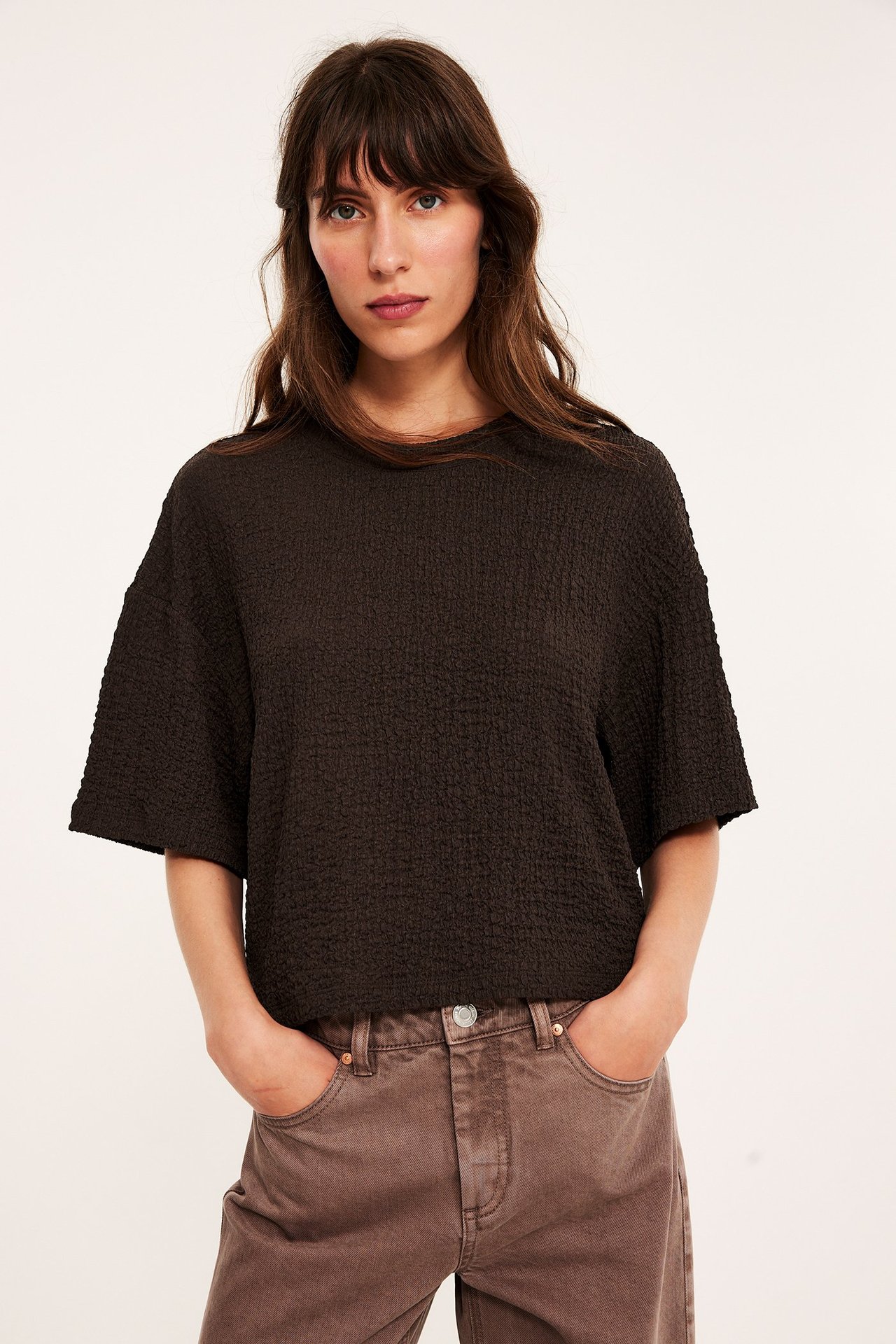 Widok z przodu kobiety w ciemnobrązowym, teksturowanym, oversize'owym T-shircie i brązowych spodniach.