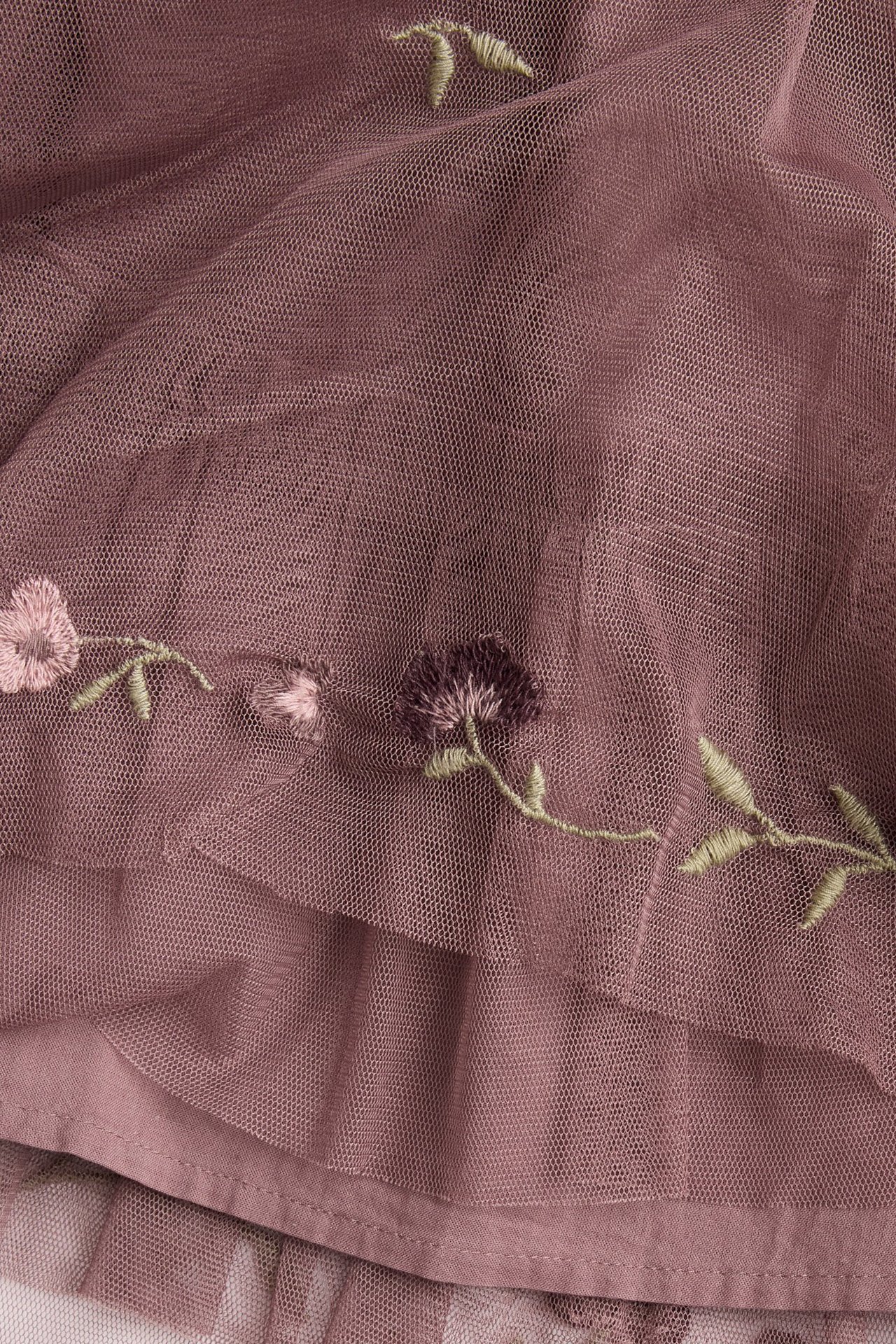 Nærbilde av en mørk rosa meshkjole med broderte rosa og lilla blomster og grønne blader.