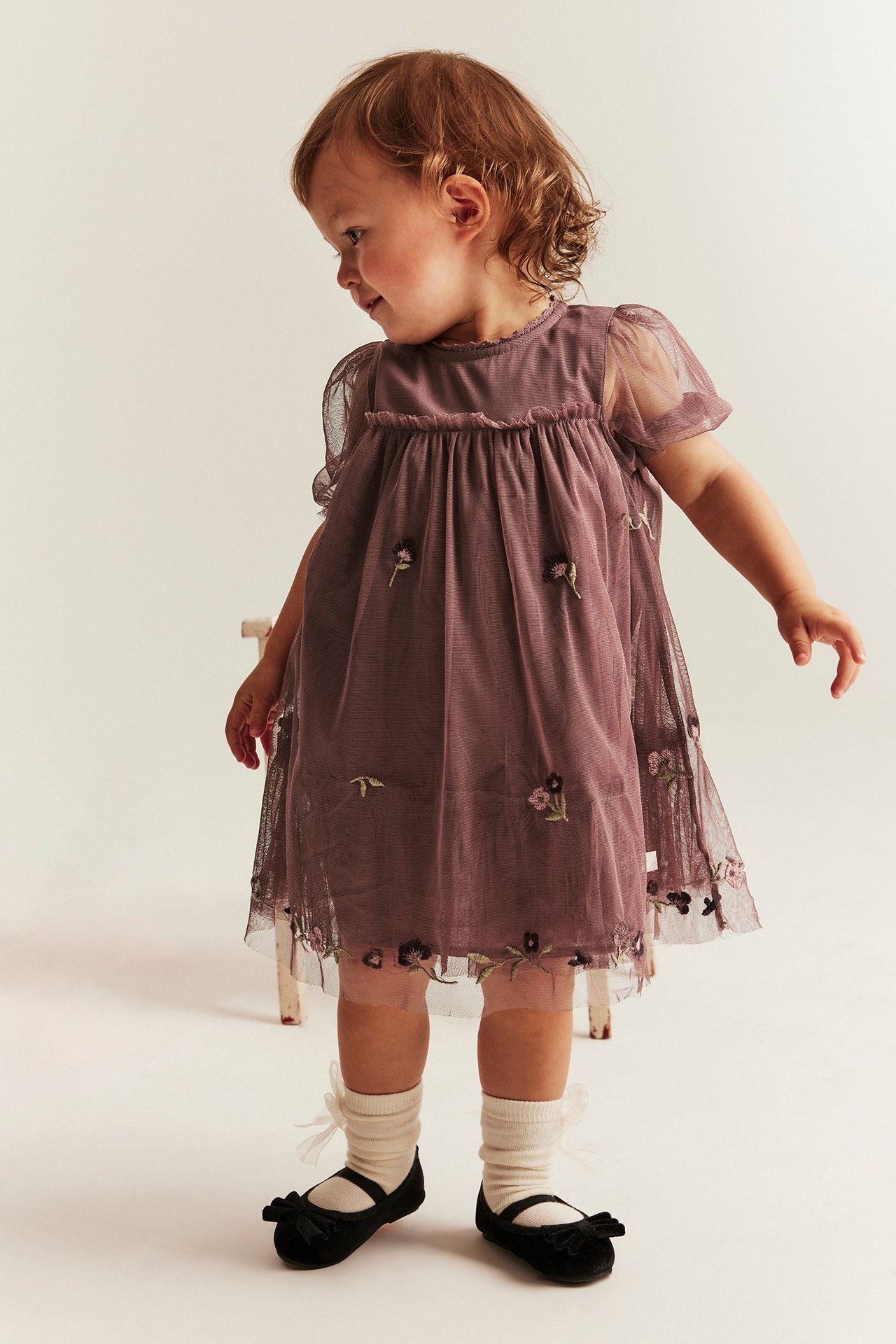 Frontvy av baby i mörklila meshklänning med broderier, puffärmar, gräddvita strumpor och svarta ballerinaskor.