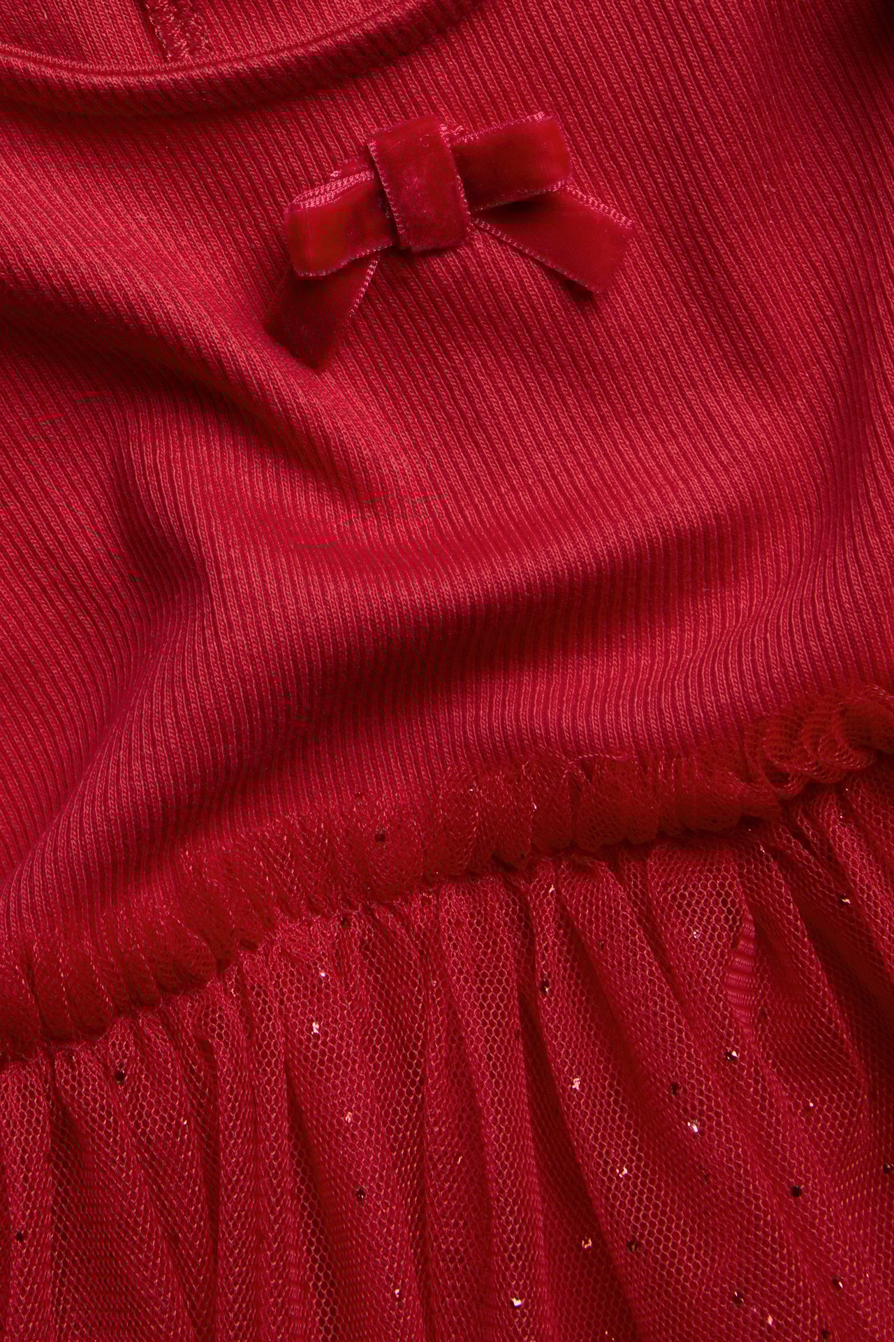 Nærbilde av rød babykjole med ribbestrikket topp, fløyelsløyfe og glitrende netting skjørt.